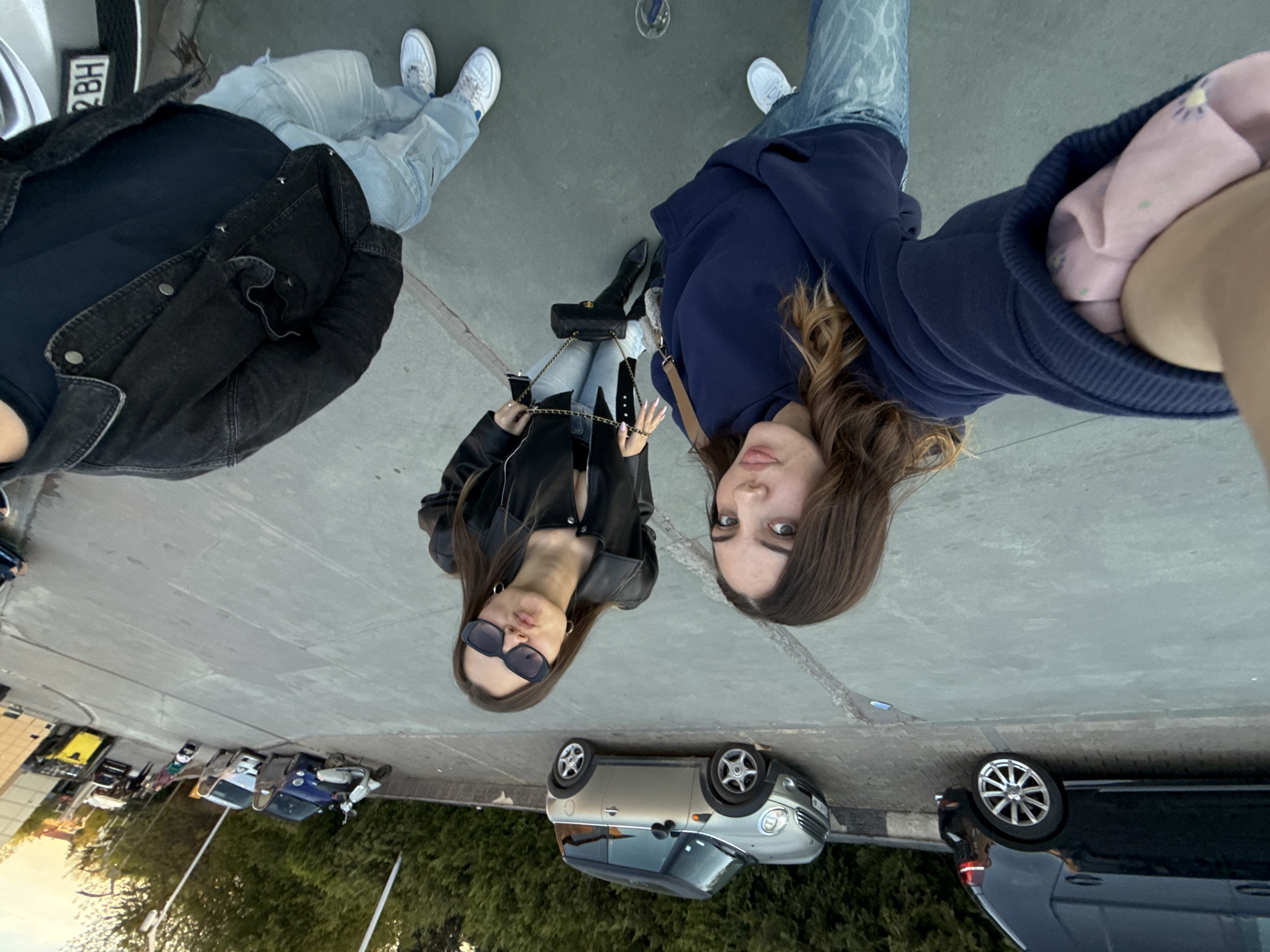 The image shows a selfie taken by a person wearing a blue hoodie, with two other women present. One woman is wearing a black leather jacket and sunglasses, while the third is wearing a black denim jacket. All three are standing on a paved surface, presumably a sidewalk. Several parked cars are visible in the background, suggesting an urban setting. Based on the cars and the general environment, the location is likely in Sofia, Bulgaria.