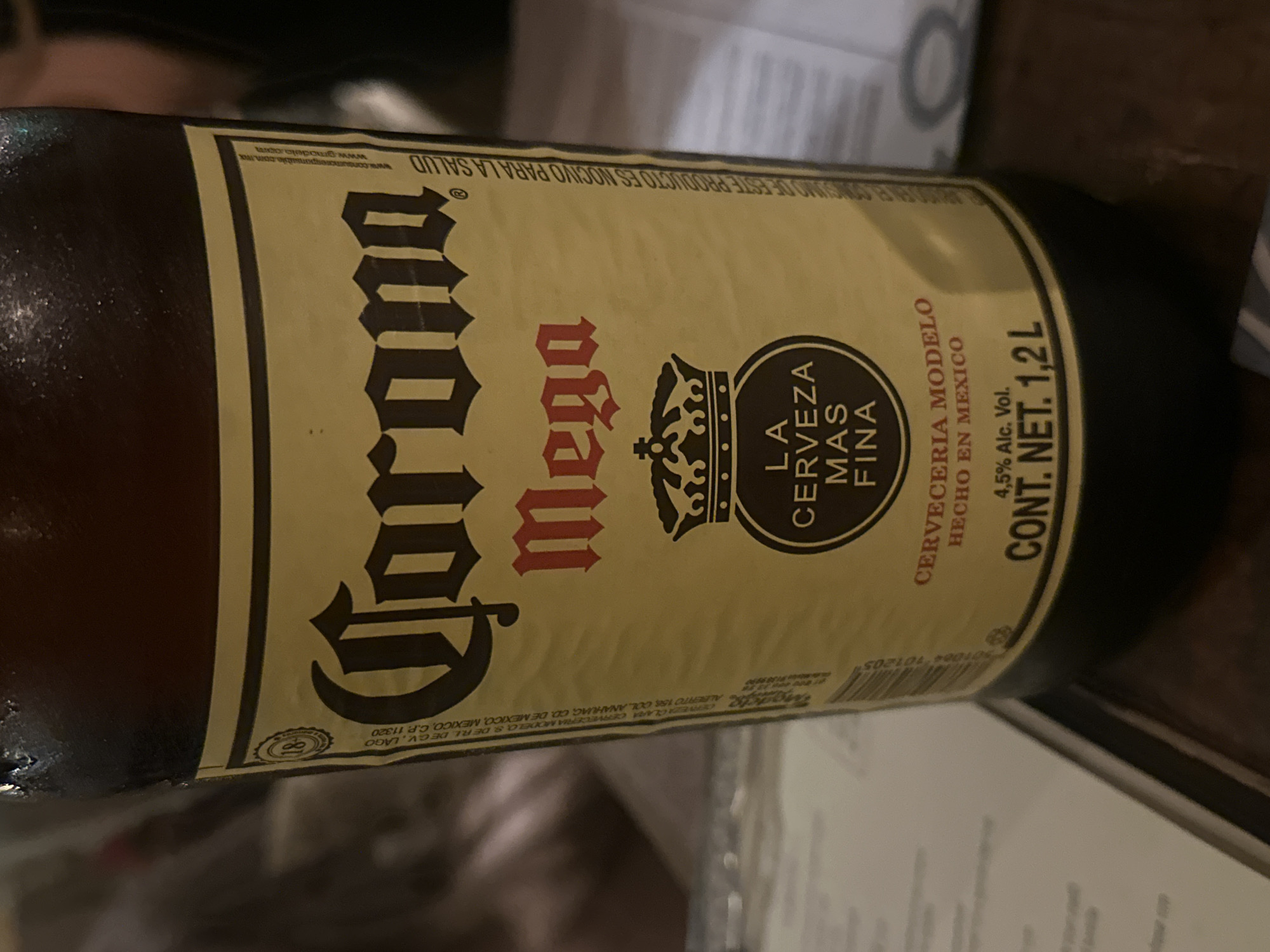The image shows a close-up of a brown glass bottle of Corona Mega beer. The label is predominantly tan with black lettering for "Corona" and red lettering for "Mega". The label also includes a crown logo and text that reads "LA CERVEZA MAS FINA". Other label details include "CERVECERIA MODELO", "HECHO EN MEXICO", and "CONT. NET. 1,2 L". The label also lists the beer's alcohol content as "4,5% Alc. Vol." There are additional lines of smaller print on the label, including website addresses and legal information. The background is blurry, revealing a glimpse of a white surface and a dark, indistinct object. The label indicates the product is from Mexico.