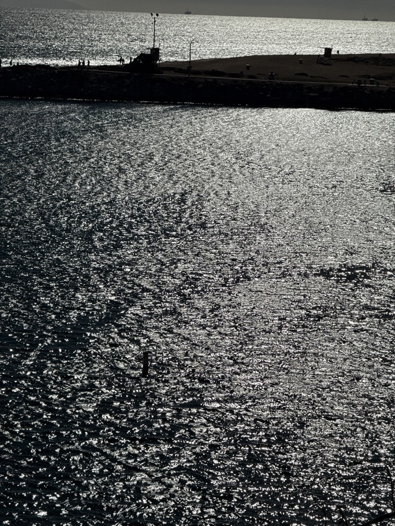 The image depicts a glistening body of water, likely the ocean or a bay, under bright sunlight, with a landmass visible in the background. The water surface is covered in light reflecting the sun, creating a sparkly effect. The landmass appears to be a breakwater or a strip of land bordering the water. Silhouetted figures and structures are visible on the land, including what seems to be a lifeguard tower.
Based on the presence of a breakwater, lifeguard tower, and the coastal landscape, this image is likely from Newport Beach, California, United States.
