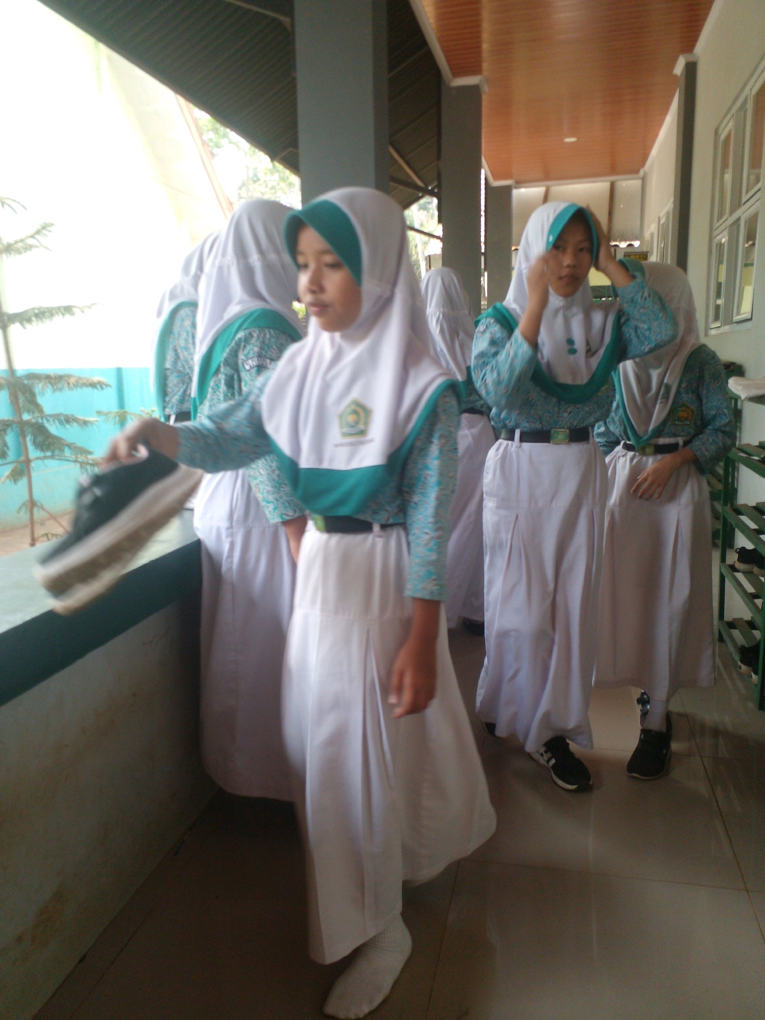 In Banyuputih, Indonesia, an outdoor scene depicts several young females in school uniforms. One girl in the foreground, wearing a white hijab with a teal accent and a matching teal and white patterned blouse and white skirt, holds up a black and white sneaker in her right hand. She appears to be in motion. Behind her, other girls in similar uniforms stand, their faces partially obscured. The environment features a walkway with a tiled floor and a railing. The lighting suggests it is daytime, with sunlight filtering from the left. A green railing is visible on the right side of the frame.