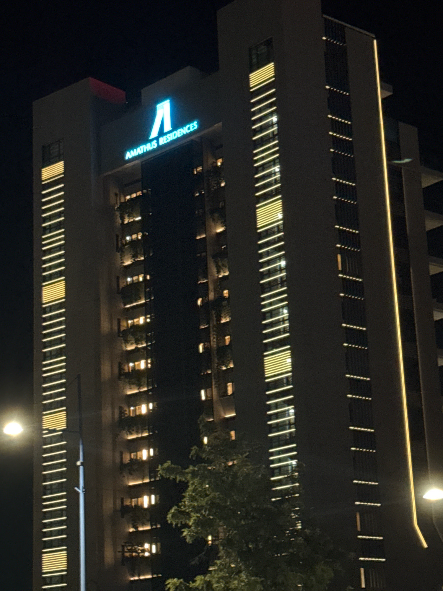 A nighttime, eye-level, full shot captures the exterior of the Amathus Residences building in Agios Tychona Community, Cyprus. The modern, tall building is illuminated by a combination of exterior lighting and interior lights visible through windows. A prominent blue sign at the top of the building reads "AMATHUS RESIDENCES" with a geometric symbol above it. The facade features vertical bands of yellow light on the left and smaller, scattered lights on the right. Trees are partially visible in the foreground, obscuring the lower portion of the building and some of its lighting. The sky is dark, indicating nighttime.