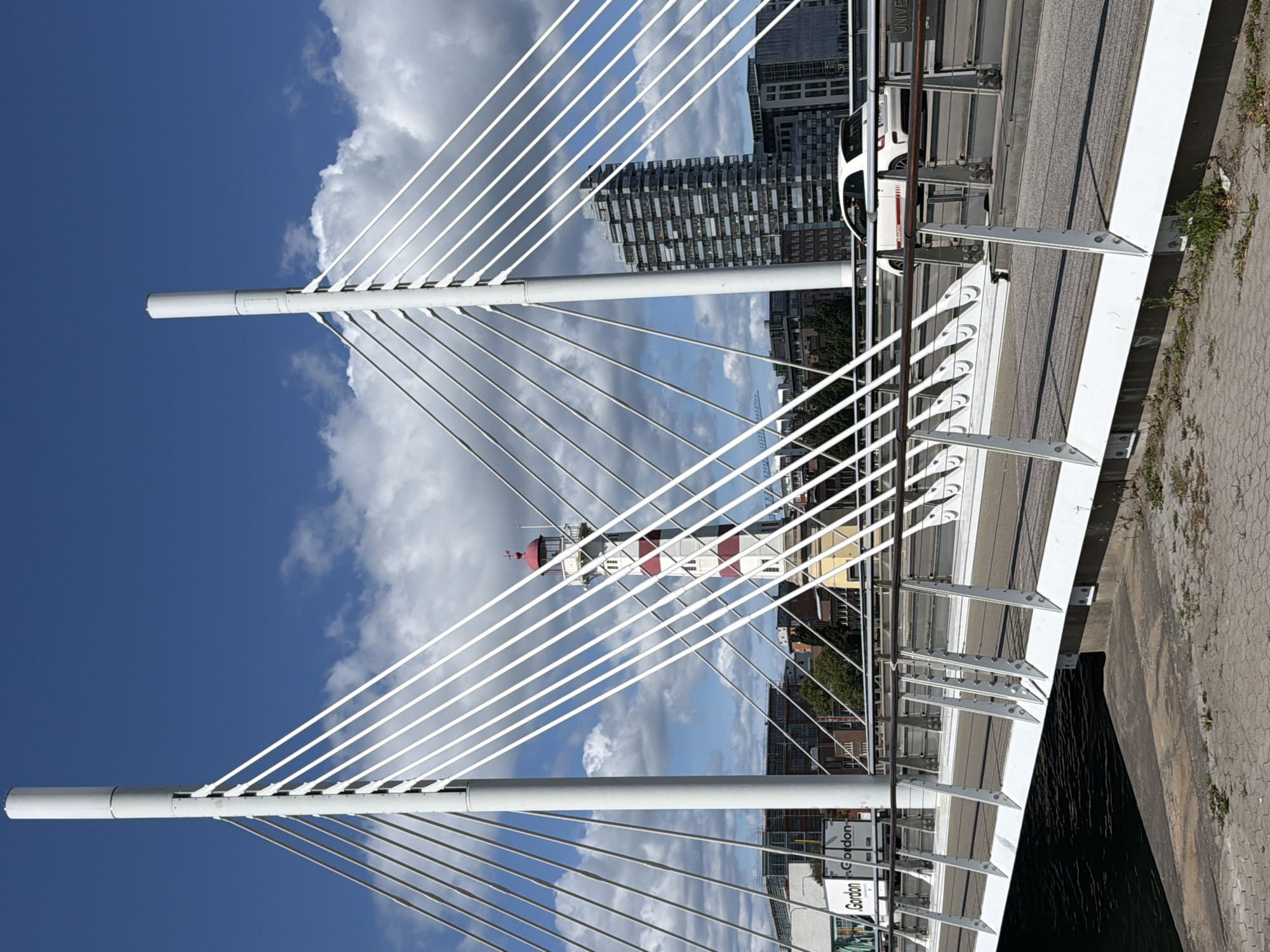 The image depicts the Kockumskajen bridge in Malmö, Sweden, a modern cable-stayed bridge characterized by its white support towers and numerous thin cables. The bridge spans a body of water, with a concrete walkway and road surface visible. A white Fiat 500 Abarth is shown driving on the bridge. In the background, a distinctive red and white striped lighthouse stands prominently. Behind the lighthouse, a tall, modern apartment building with a grid-like facade dominates the skyline. Other buildings and trees are also visible in the urban landscape under a bright blue sky with scattered white clouds. The foreground shows a paved walkway along the water's edge.