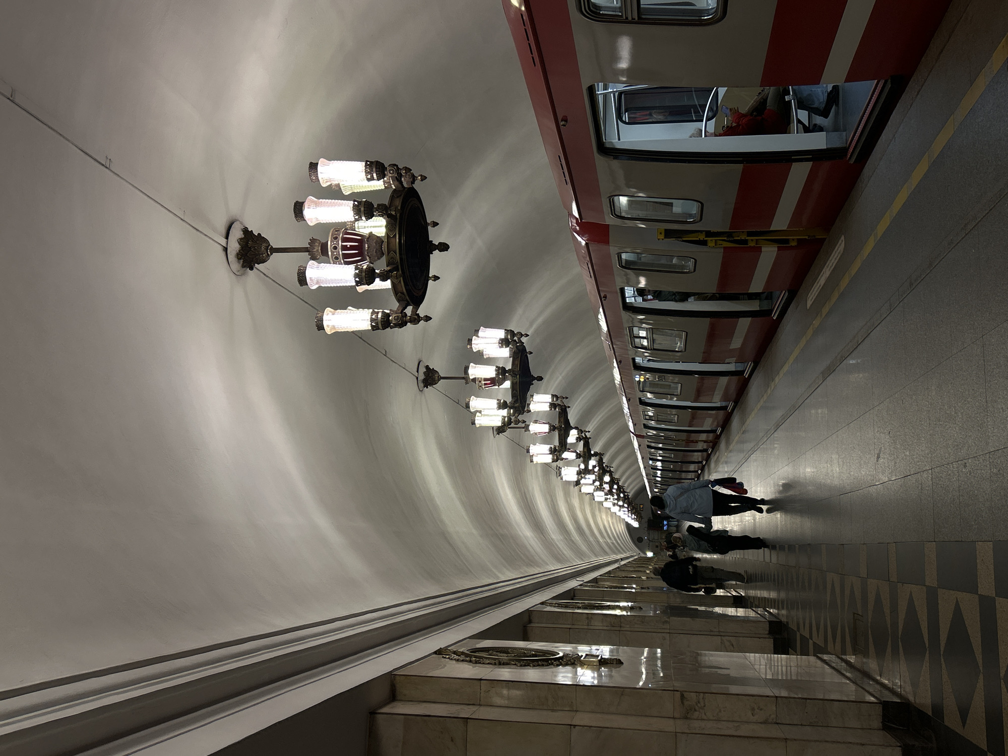 The image shows a platform in a Saint Petersburg Metro station. A red and white train is stopped at the platform. Several people are walking along the platform. Ornate chandeliers hang from the arched ceiling. The platform is tiled.
