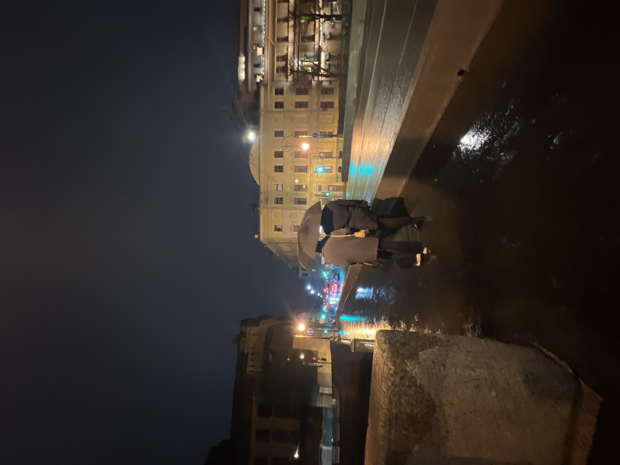 The image shows a nighttime scene in Saint Petersburg, Russia. Two people are walking under an umbrella on a wet sidewalk next to a body of water, likely a canal or the Neva River. The street and sidewalks are shiny with rain, reflecting city lights. In the background, buildings are illuminated by streetlights and windows. Traffic signals are visible. The scene suggests a rainy evening in the city center.
