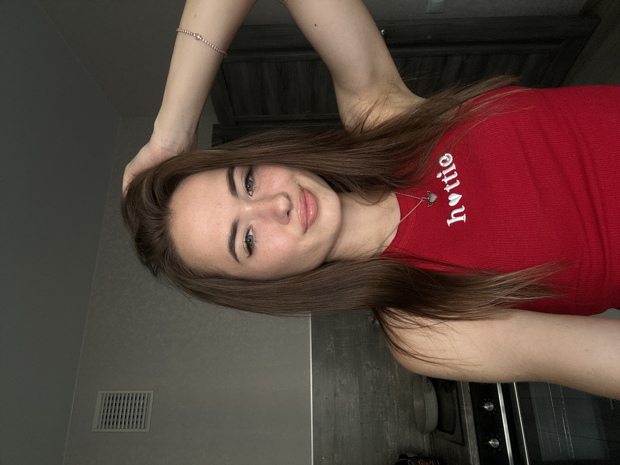 A young woman with long, brown hair is captured in a selfie-style shot, looking directly at the camera. She is wearing a red ribbed tank top with white lettering that reads "hottie" and a small, silver necklace with a heart pendant. Her left arm is raised, and she is holding her hair with her hand. She has light skin, appears to be wearing makeup, and has a subtle smile. The background suggests an indoor setting, possibly a kitchen, with a gray wall and a vent visible on the upper left. A stovetop and oven are partially visible to the left, and a dark door is to the right. The lighting is dim.