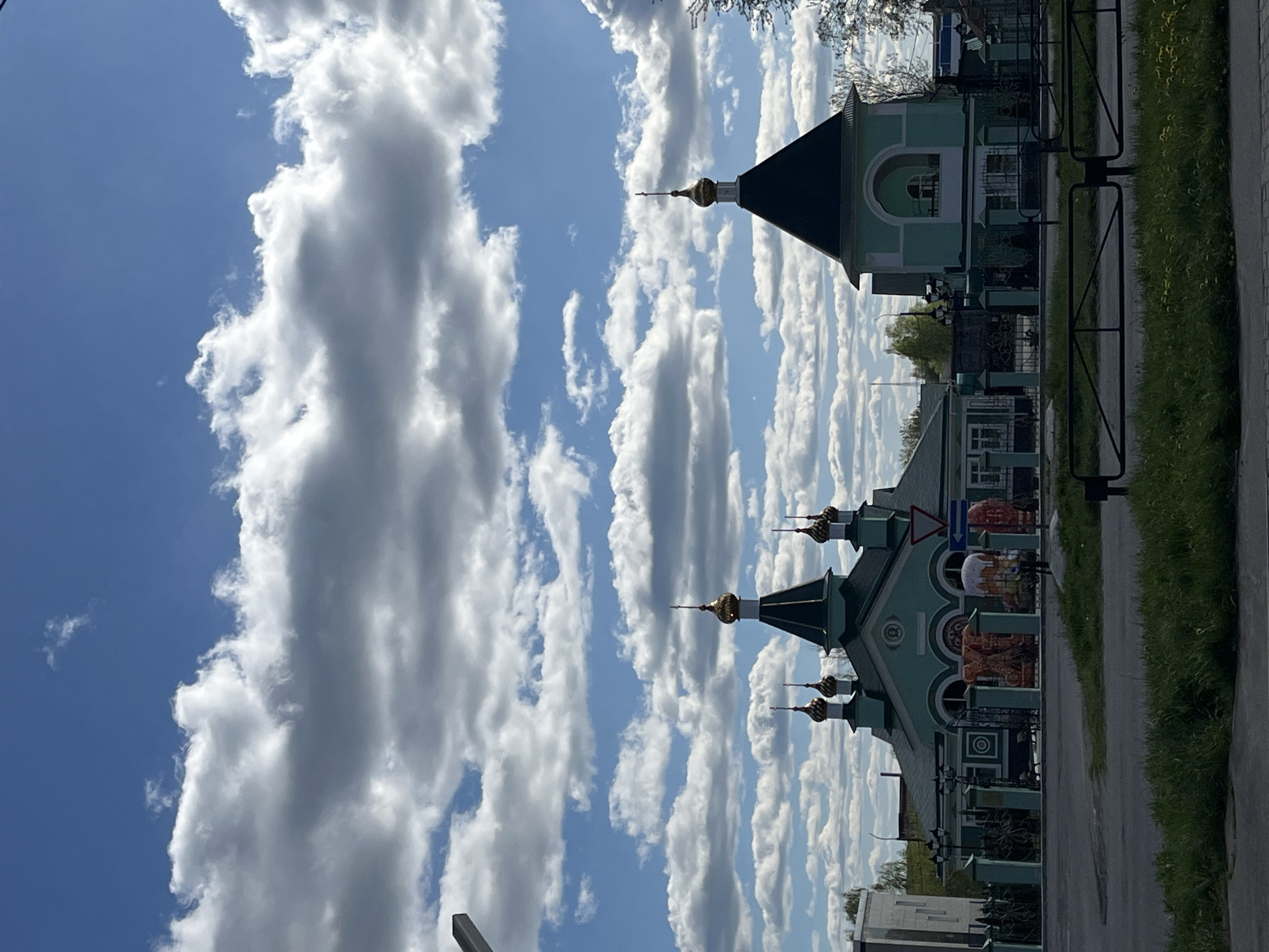 The image shows a building with distinct architectural features, including multiple domes topped with golden crosses, painted in shades of light green. The building is situated alongside a paved sidewalk, bordered by a wrought iron fence and green vegetation, likely grass. A few people are visible near the building. The sky is blue with numerous elongated white clouds. The location is Chaikovsky, Russia.