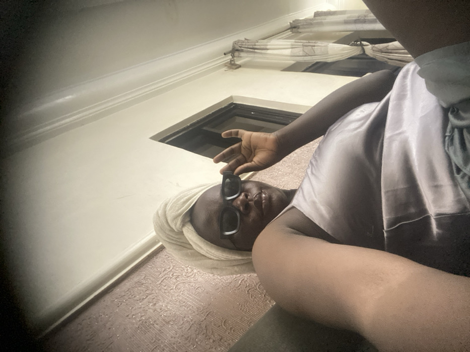 An individual with dark skin wears a towel on their head and sunglasses. The person lies on a textured surface with the head tilted towards the left, near a window. A partially visible arm reaches up, fingers near the sunglasses. The person wears a satin or silk top. White walls surround the scene. Curtains and molding add detail. The overall image is taken from a low angle, looking upward.