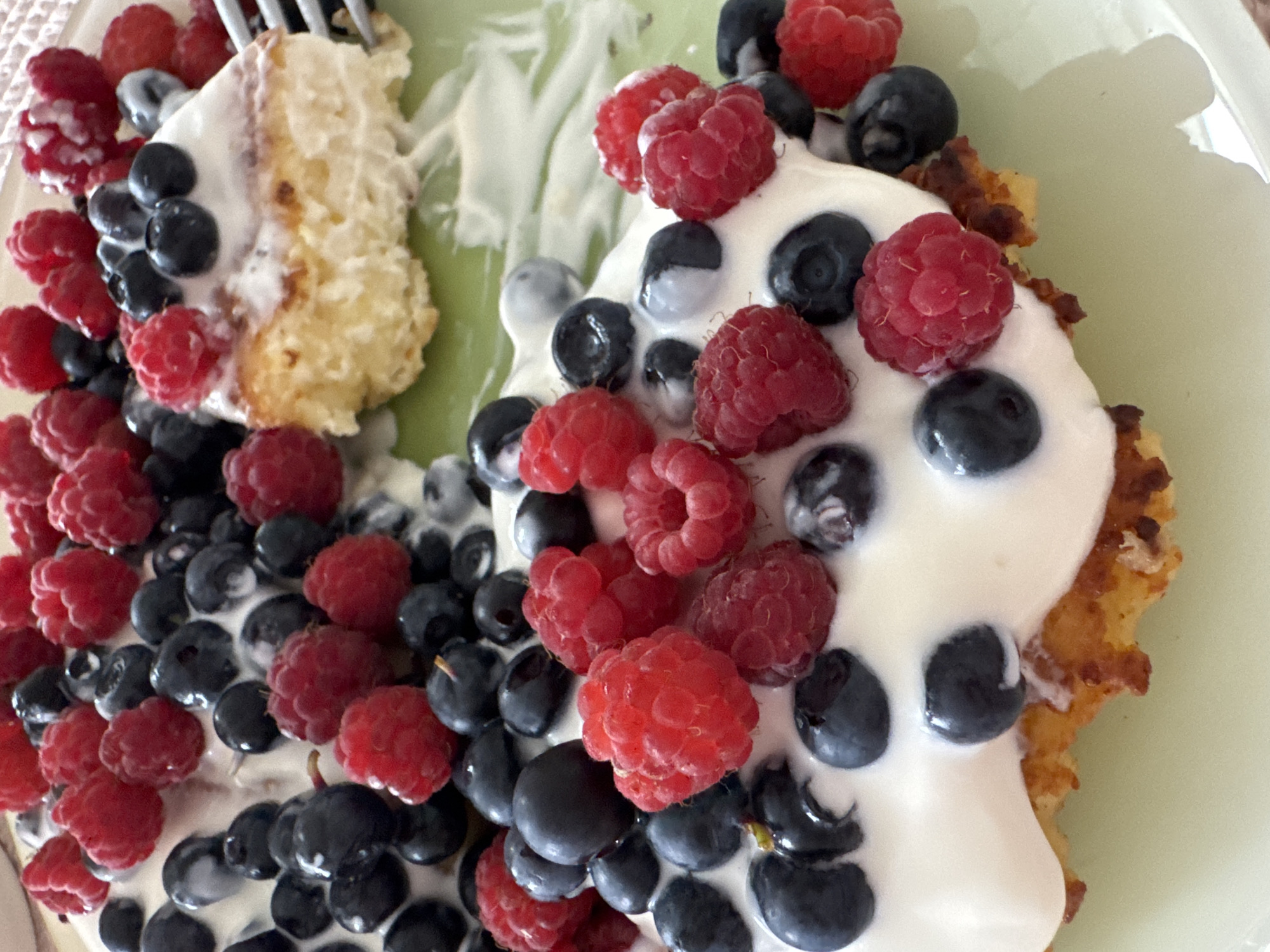A close-up, overhead shot captures a plate of food featuring a golden-brown waffle topped with a generous swirl of white cream and an abundance of fresh raspberries and blueberries. A slice has been removed from the waffle, revealing its fluffy interior. The food rests on a pale green plate. The image is cropped to focus tightly on the food, with the background blurred and indistinct. No specific location within Ladozhskoe Ozero, Russia can be determined from the visual information provided.