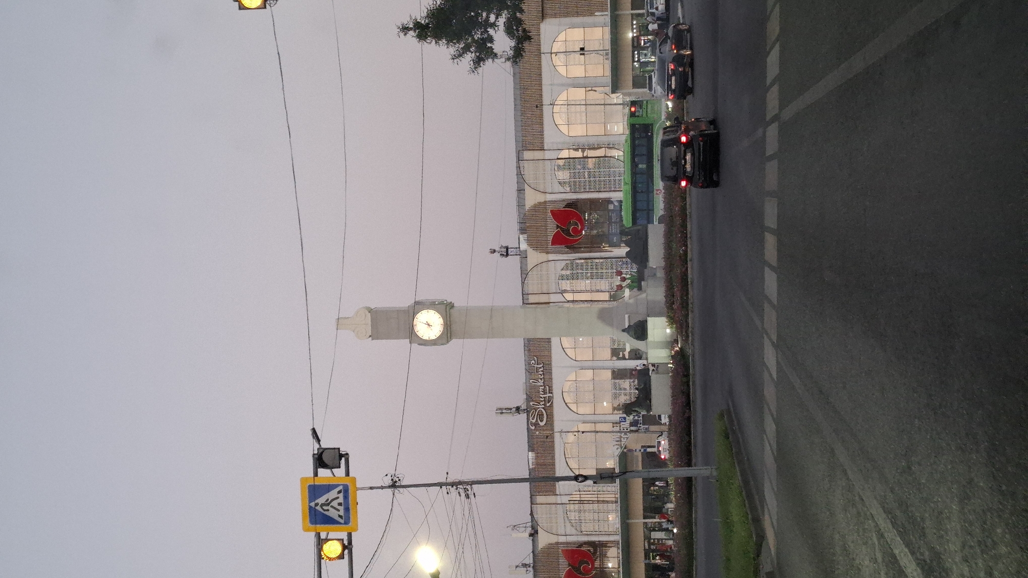 The image captures a street scene in Shymkent, Kazakhstan, focusing on the city's prominent clock tower and the building behind it, which appears to be a market or a commercial center. The sky is overcast, suggesting either dawn, dusk, or a heavily clouded day.
In the foreground, a street with a pedestrian crossing is visible. Several cars are on the road, including a black SUV in the middle lane and a black sedan to its right. Traffic lights are present, with a yellow light illuminated on the left side of the frame. A pedestrian crossing sign is also visible, accompanied by a yellow signal light.
Dominating the mid-ground is a tall, slender clock tower with a visible clock face displaying a time that appears to be approximately 10:05. The tower is positioned in front of a large building with a distinctive architectural style, featuring arched windows and the word "Shymkent" inscribed in script above the entrance. The building is decorated with large, red floral motifs. A green bus is seen driving past the building.
The lower portion of the scene includes a flowerbed with pink and red flowers, and a green lawn strip to the left. In the background, some smaller structures and trees are discernible. The overall impression is that of an urban environment, likely a central square or intersection in Shymkent, given the prominent signage.