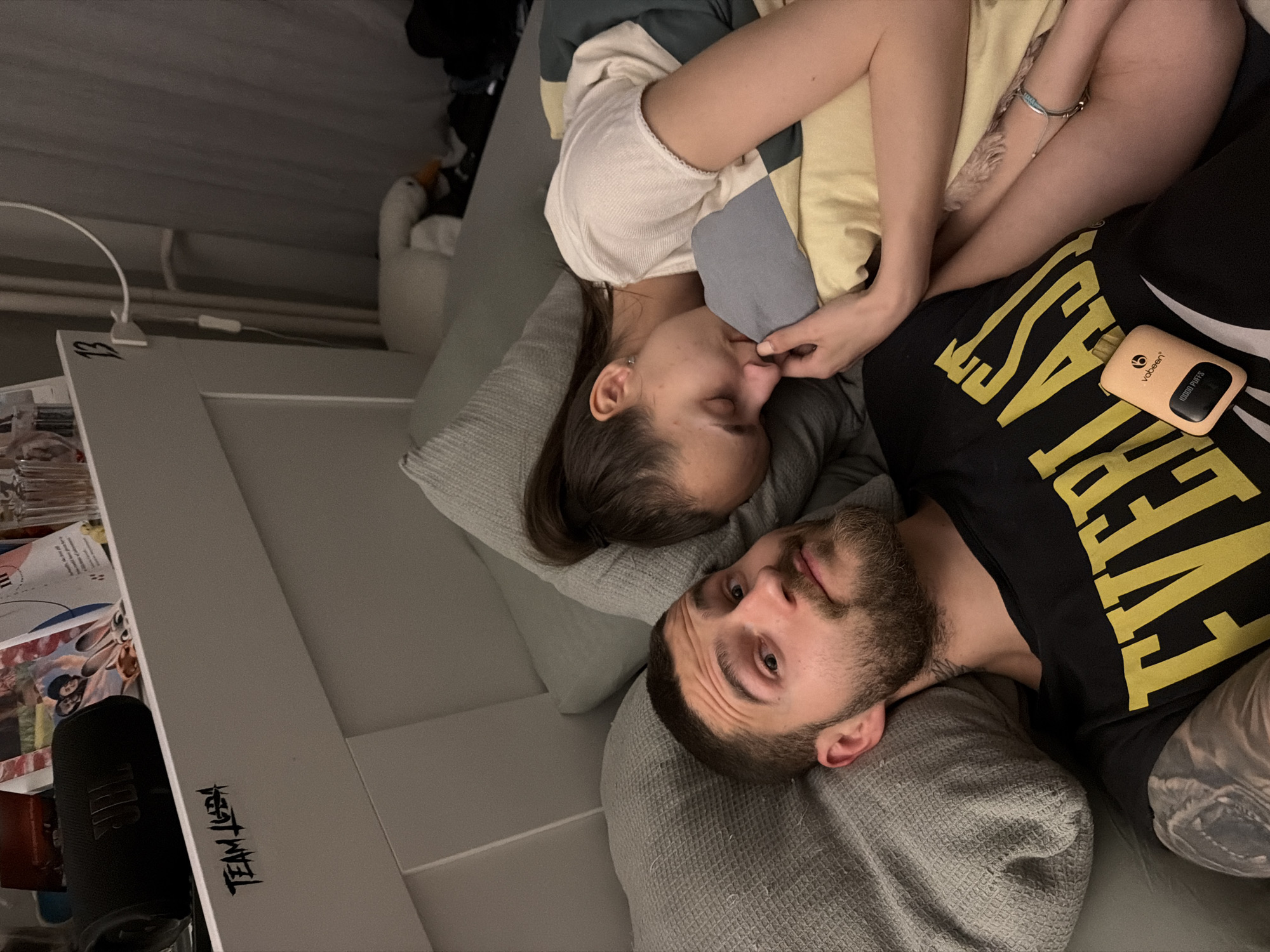 An overhead shot captures two individuals resting indoors, likely in a bedroom or living area in Sofia, Bulgaria. The image is rotated counter-clockwise.
A male-presenting individual, with a beard and short dark hair, lies on his back on a grey textured pillow, looking directly at the camera. He wears a black t-shirt with yellow text, partially visible as "EVERLAST". Tattoos are visible on his arm and neck. Next to him, a female-presenting individual lies on her side, facing away from the camera, with her eyes closed, suggesting she is sleeping or resting deeply. Her dark hair is pulled back, and she wears a light-colored top, snuggled into a patterned duvet or blanket (yellow, blue, cream). A thin bracelet is on her wrist, and tattoos are visible on her arm.
The overall lighting is dim, indicating it might be evening, night, or simply low indoor light. The atmosphere appears peaceful and relaxed.
Several objects are scattered around them. On the male's side, an orange/beige vaping device is visible, bearing the brand "vabeen" and the text "10000 PUFFS". A white plush toy resembling a goose or duck peeks from the background near the female. To the left, a light-colored shelf or bedside table holds various papers/magazines, a black JBL speaker, and a white charging cable. The number "13" is written in black on the edge of this surface, and the stylized text "TEAM ZED" is also visible.