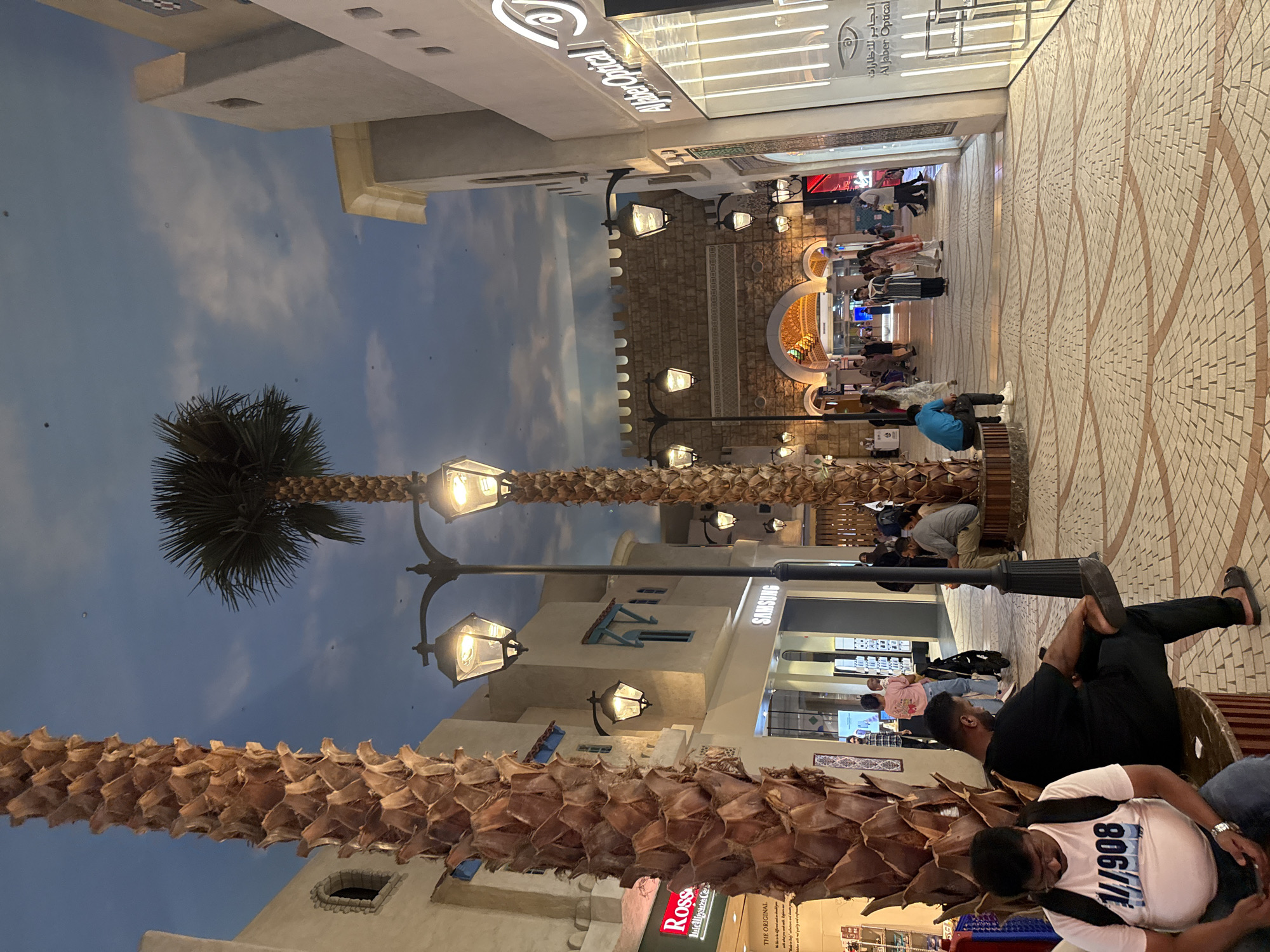 The image depicts an indoor marketplace in Dubai, United Arab Emirates, designed to resemble an Arabian streetscape. Two tall, dried palm trees dominate the foreground. The architecture features light-colored buildings with arched windows and decorative battlements, suggesting a traditional Middle Eastern aesthetic. A blue, cloudy sky is painted on the ceiling, creating an illusion of an open-air environment.
Several individuals are present, walking and sitting. In the foreground, two men are seated on a bench. The man on the left wears a light pink t-shirt with text, while the man on the right is dressed in black. Further down the marketplace, other people are seen strolling, with a variety of clothing suggesting a mix of casual and traditional attire. The ground is paved with a patterned brickwork.
Various commercial establishments are visible, including a store labeled "SAMSUNG" and another identified as "Al Jaber Optical." Street lamps with a classic design are mounted on poles, illuminating the area. The overall impression is a vibrant, pedestrian-friendly shopping or entertainment venue.