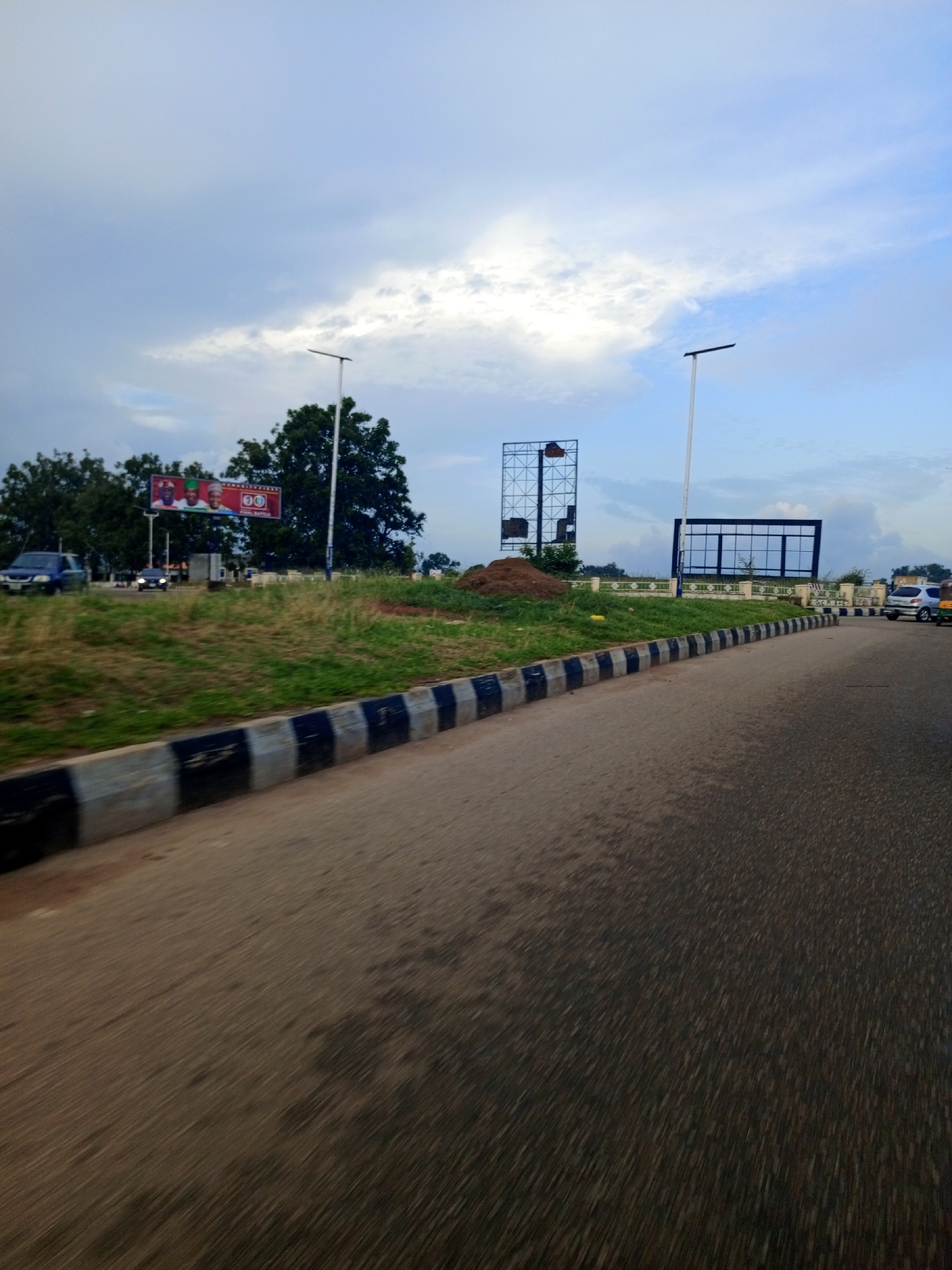The image depicts a view from a moving vehicle on a road in Jos, Nigeria. The road is paved and curves to the right. On the left side of the road, there is a grassy embankment with a curb made of alternating black and gray concrete blocks. Atop the embankment, there are several trees, some with lush green foliage. A large billboard with three portraits of men in traditional attire is visible, along with text in what appears to be English and possibly another language. Further along the embankment, there are two large, empty advertising frames, one taller and rectangular, and the other wider and rectangular with a blue border. A single street lamp with a dark angled arm stands behind the first billboard, and another similar street lamp is visible further down the road. There is a pile of dirt or sand on the embankment near the advertising frames. A dark blue car is parked on the side of the road to the left, and a few other vehicles, including a white car, are seen further down the road, suggesting traffic. The sky is overcast with dramatic clouds, hinting at either dawn, dusk, or an impending storm. The overall scene is one of a typical urban roadside environment in Nigeria.
