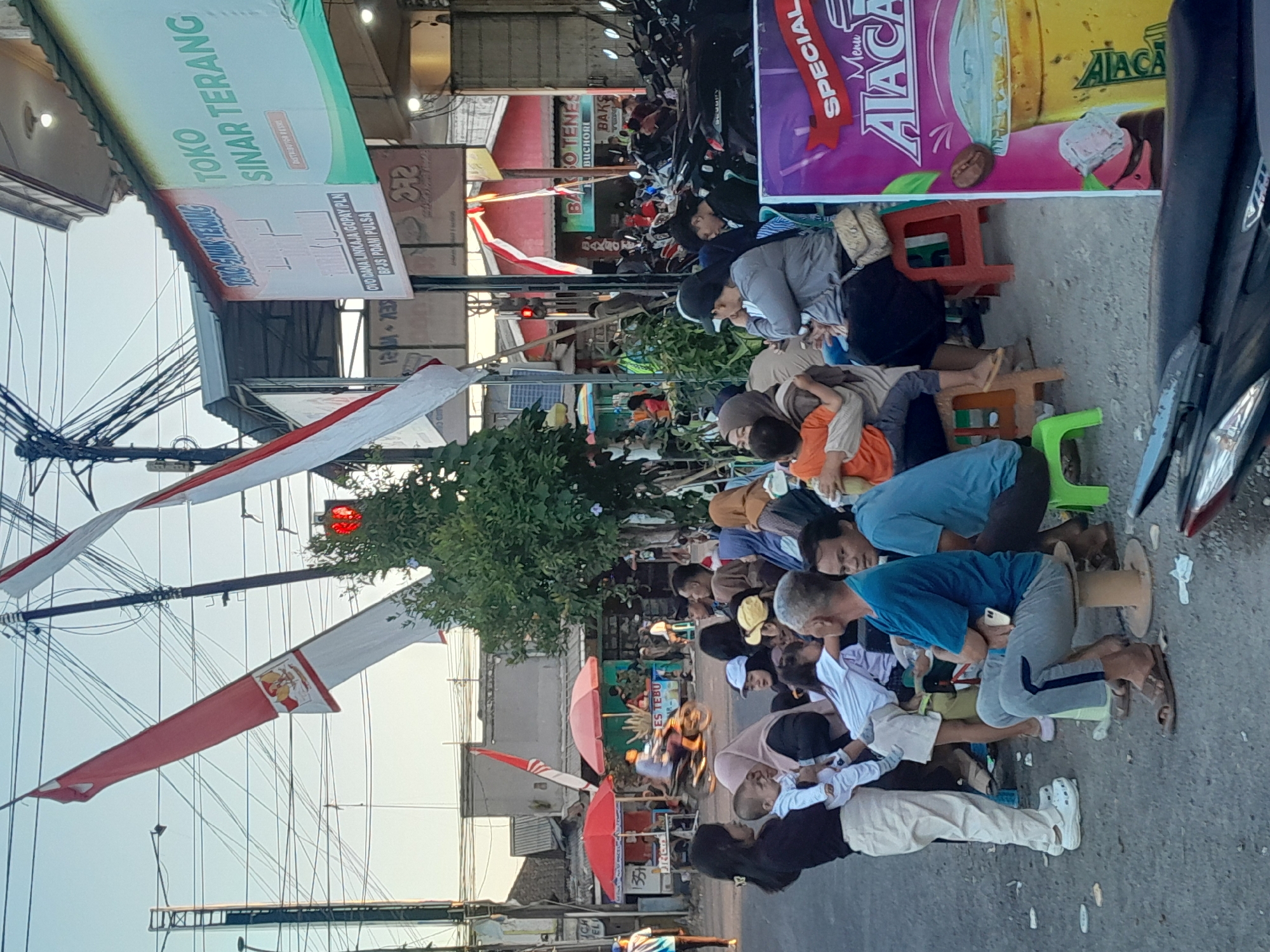 The image depicts a street scene in Gentengwetan, Indonesia, likely during the late afternoon, with a gathering of people and various commercial activities. The central focus is a group of individuals, including adults and children, sitting and standing on the side of a street, seemingly waiting or socializing. Several people are holding or interacting with young children.
Dominating the background are numerous overhead electrical wires, indicative of an urban environment. Prominently displayed are two large Indonesian flags, red and white, unfurled and suspended from poles. Adjacent to these are signs for businesses. One sign, in green, reads "TOKO SINAR TERANG," which translates to "Bright Light Store," and mentions "DISTRIBUTOR RESMI." Another sign above it, partially obscured, appears to be related to electrical services or products. Further down the street, a red traffic light is visible, suggesting the presence of an intersection.
The ground is paved with asphalt, with some scattered debris and white paper. To the left, two red umbrella-covered stalls are set up, one with a sign mentioning "ES TEBU," meaning sugarcane juice. A motorcycle is seen passing by in front of these stalls, with a rider in a yellow shirt.
Further to the right, a row of parked motorcycles lines the street. A large, colorful advertisement poster is placed near the group of people, featuring text like "SPECIAL Menu" and an illustration of a beverage. The architectural style of the buildings in the background suggests a mix of commercial and residential structures. The overall atmosphere appears to be a bustling local street with a blend of everyday life and commerce.