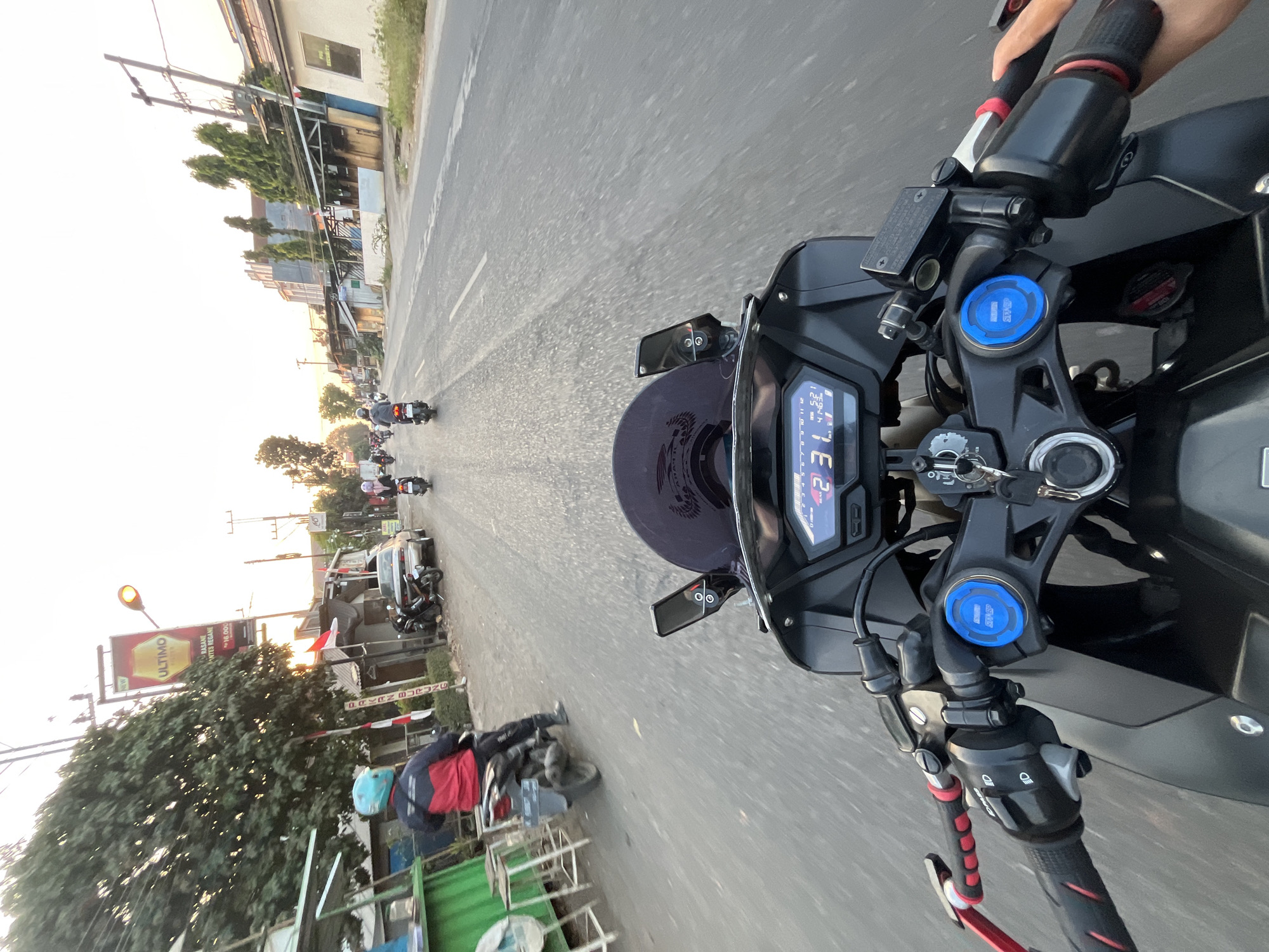 The image captures a first-person perspective of someone riding a motorcycle on a street in Pelang, Indonesia. The rider's hands are visible on the handlebars, and the motorcycle's dashboard displays speed and other information. In the background, other motorcycles and cars are present on the road, along with buildings and trees lining the street. A large banner with text and an image of a bottle can be seen on the left side of the road, likely a billboard. The sky suggests it is either early morning or late afternoon, with a warm, hazy light. The overall scene depicts a typical urban street environment in Indonesia during daylight.