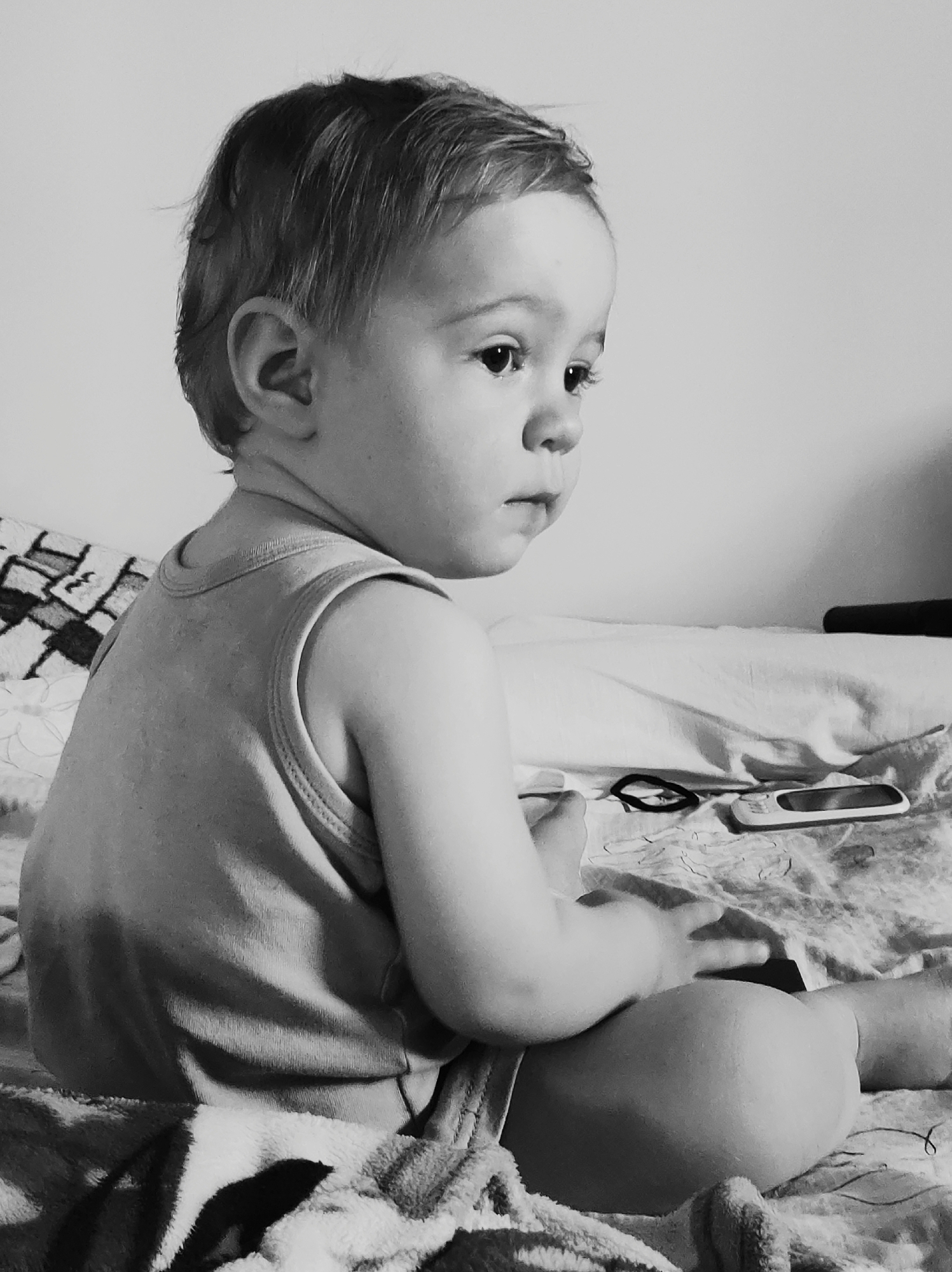 A black and white, medium shot photograph depicts a baby sitting on a bed. The baby is facing away from the viewer, looking to the right of the frame. The baby is wearing a sleeveless shirt. The bed has a patterned blanket and a white sheet. A phone and a hair tie are on the bed next to the baby. The background is a plain, light-colored wall. The location and city are Unknown.