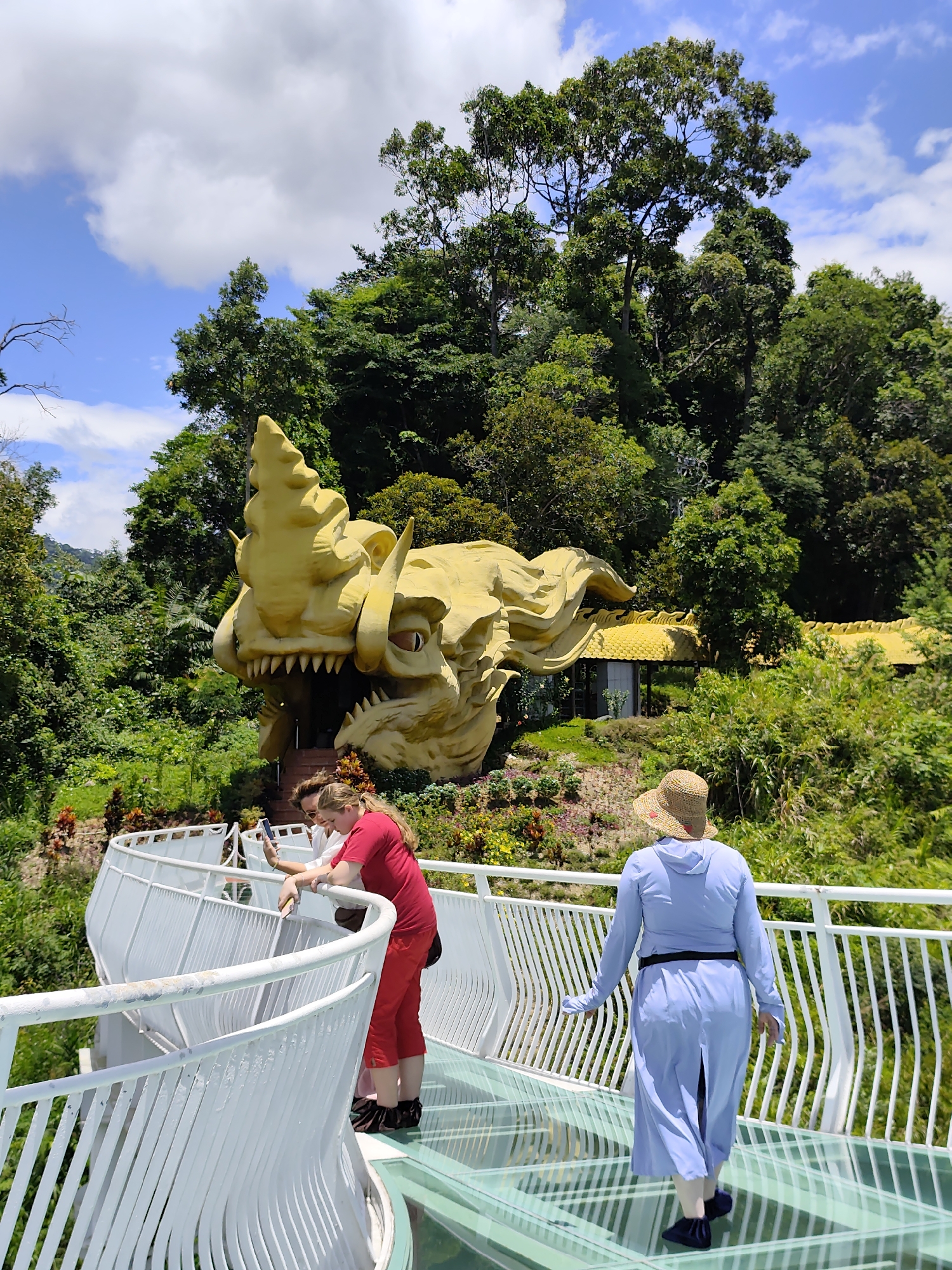 In a scenic outdoor environment with lush green foliage and a bright blue sky with scattered clouds, a large, golden dragon sculpture with sharp teeth and scales dominates the background. In the foreground, a white, curved walkway with a glass floor is occupied by two individuals. On the left, a person wearing a red shirt and red pants is looking at their phone. Beside them, another person in a light blue long dress and a straw hat is walking away from the camera. The walkway offers a view of the dragon sculpture and the surrounding tropical vegetation.