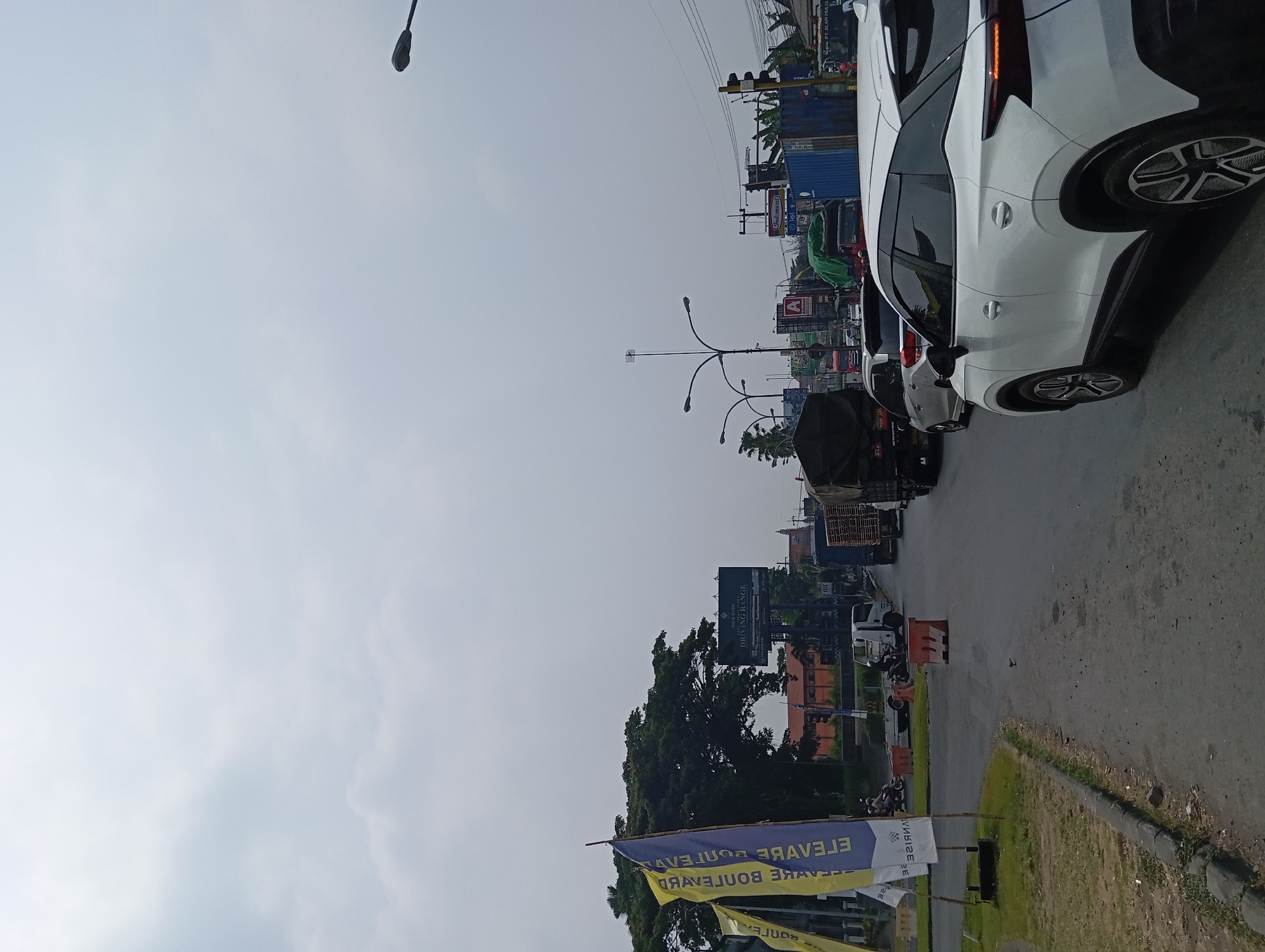 The image shows a busy street scene in Karang Jati, Indonesia, under an overcast sky. A white SUV is in the foreground, facing right, with its rear passenger side visible. Traffic is backed up on the road, with several trucks and cars ahead. In the background, there are buildings, trees, billboards, and streetlights. One billboard prominently displays "INDOMARET" and another advertises a "DRIVING RANGE." Several flags are planted on the left side of the road, with text that appears to be in a foreign language. The overall impression is one of a typical urban traffic jam.