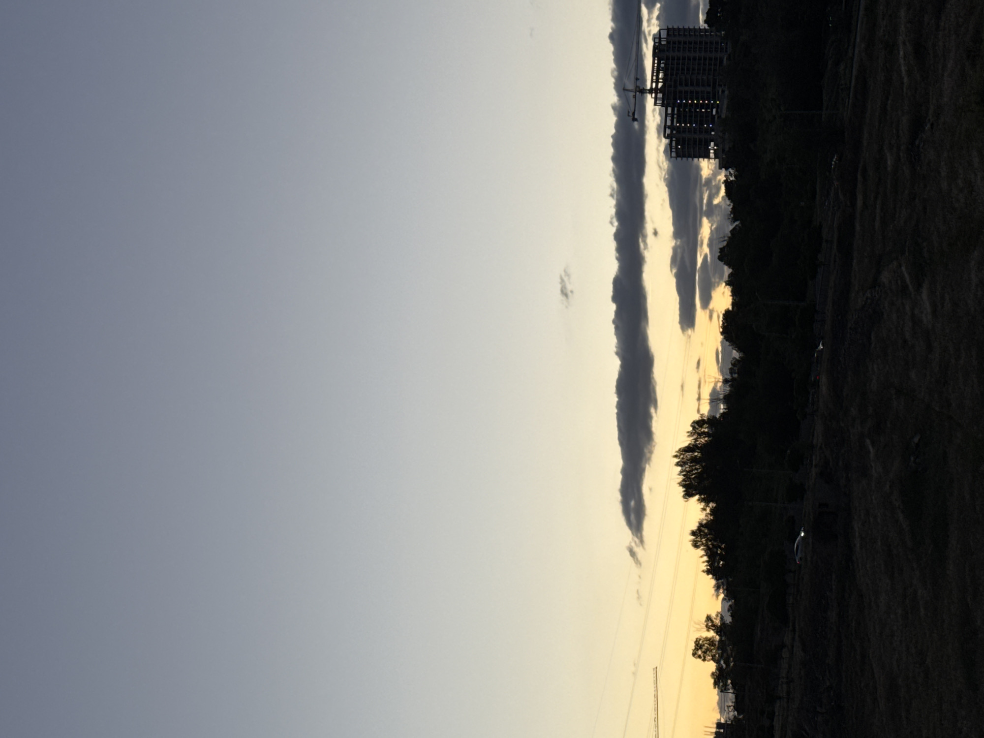 The image captures a sunset scene over what appears to be a developing area, specified as Yerevan, Armenia. The sky dominates the frame, displaying a clear gradient from a cool blue-grey in the upper left to a warm, soft golden yellow-orange at the horizon line on the right, characteristic of twilight or early evening. A prominent, elongated dark grey cloud band stretches horizontally across the middle of the sky, with several smaller clouds scattered around, some catching the warm light.
Against the brightly lit horizon, a dark, rugged landscape or hilly terrain is silhouetted on the right side of the image, bordered by a dense line of dark trees. Further to the right, the skeletal structure of a multi-story building under construction stands out as a dark silhouette, flanked by tall cranes. Faint points of light are visible within the building's framework, possibly reflections or early evening lights. In the lower right foreground, the ground is dark and uneven, suggesting undeveloped land. Subtle power lines and utility poles are visible as dark lines against the illuminated sky. A faint, light-colored car can be seen as a small silhouette on the dark ground. No discernible text is visible in the image.
