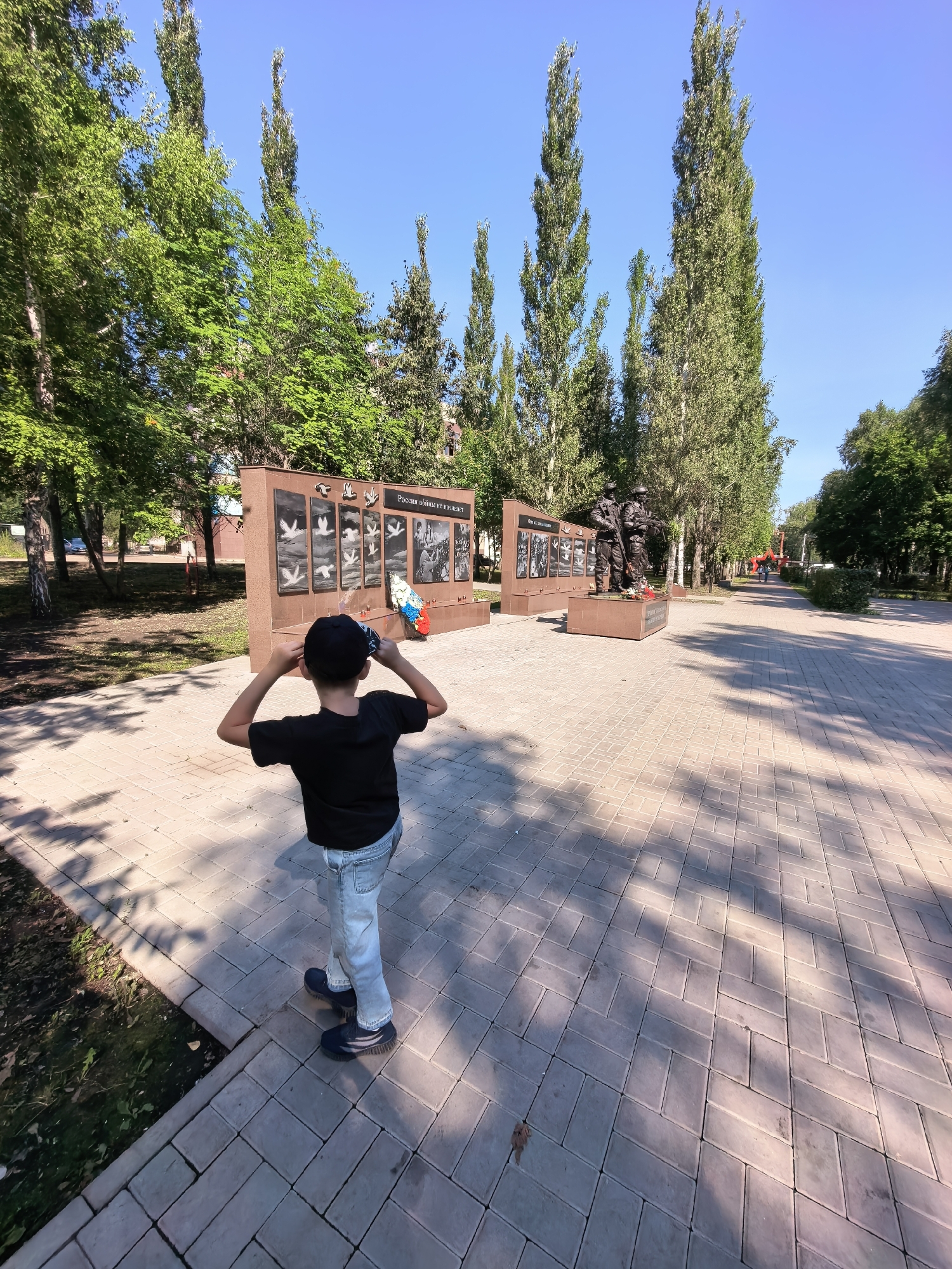 A young boy wearing a black t-shirt, blue jeans, and a black baseball cap stands with his back to the camera, looking at a war memorial in Sterlitamak, Russia. The memorial features bronze statues of soldiers and a wall with relief carvings and text. The boy is holding his hands up to his ears, as if blocking out sound. The memorial is located in a park with trees and a paved walkway. The sun is shining, casting shadows on the ground.