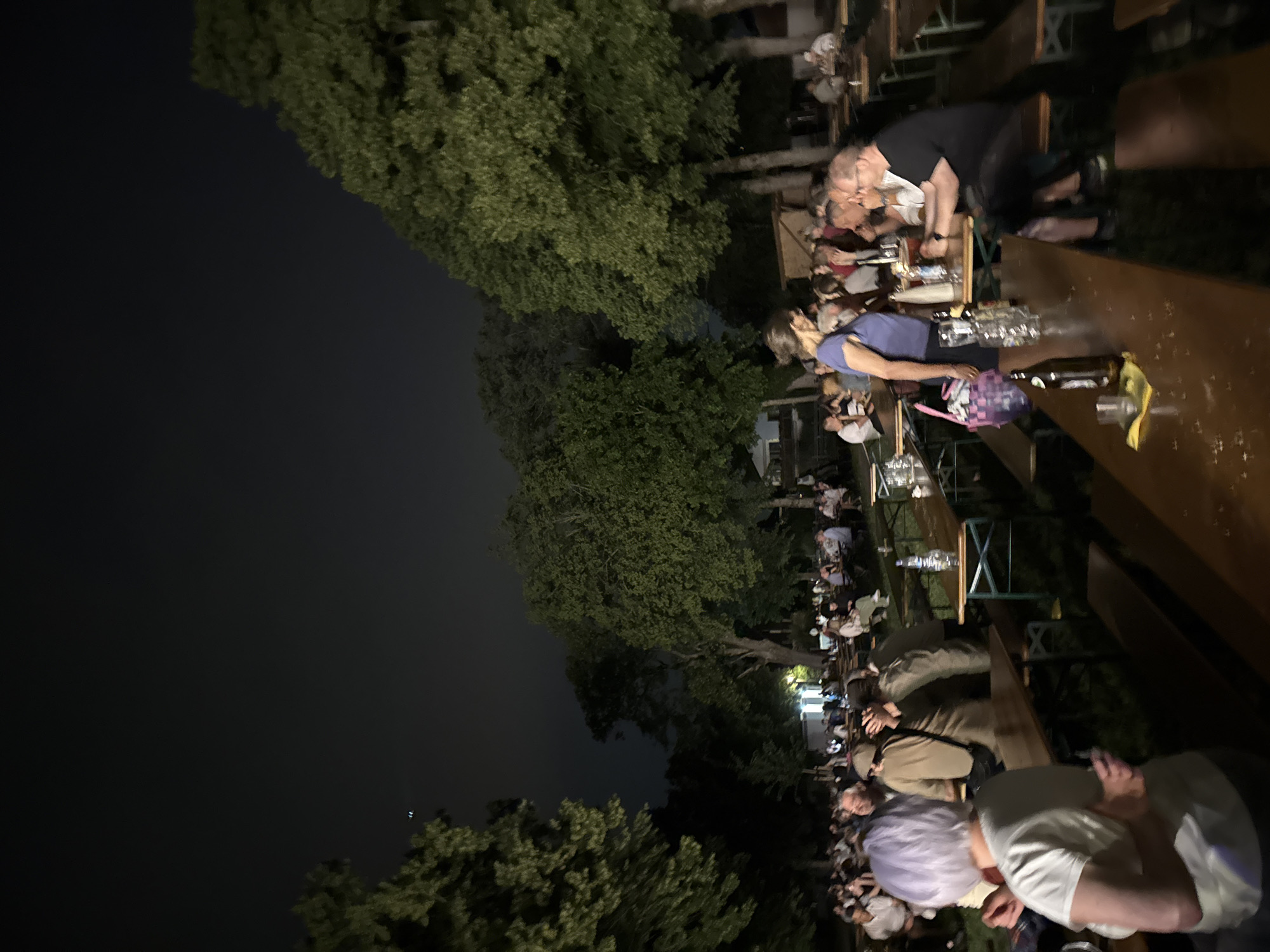This image depicts an outdoor evening gathering at the Kulturverein in Höhenkirchen-Siegertsbrunn, Germany. A large number of people are seated at long, wooden picnic tables set up outdoors, illuminated by artificial light. The atmosphere suggests a social event, possibly a beer garden or festival, with drinks visible on the tables. Lush green trees surround the area, and the sky is dark, indicating it is nighttime. The gathering appears to be a community event, with individuals of various ages engaged in conversation and enjoying the evening.