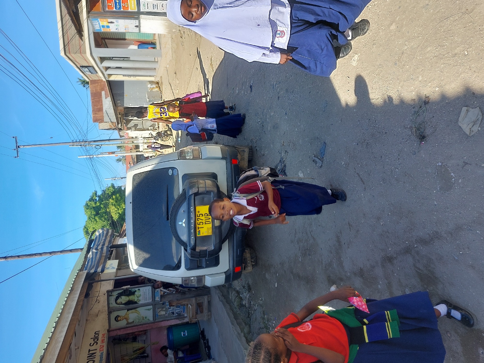 The scene depicts a street in Dar es-Salaam, Tanzania. Several children in school uniforms are present. One girl, positioned near the rear of a white Mitsubishi Pajero (license plate T575 DVR), wears a maroon and white top with a dark blue skirt. Another girl, wearing a white headscarf, is seen near the top of the frame. Another is on the bottom wearing a red shirt and blue skirt with green and yellow stripes. A "POINT Salon" is visible on the left, with its signage and contact number. Overhead electrical wires crisscross the blue sky. Buildings line the street, and a utility pole is present on the left side. The street surface appears paved.