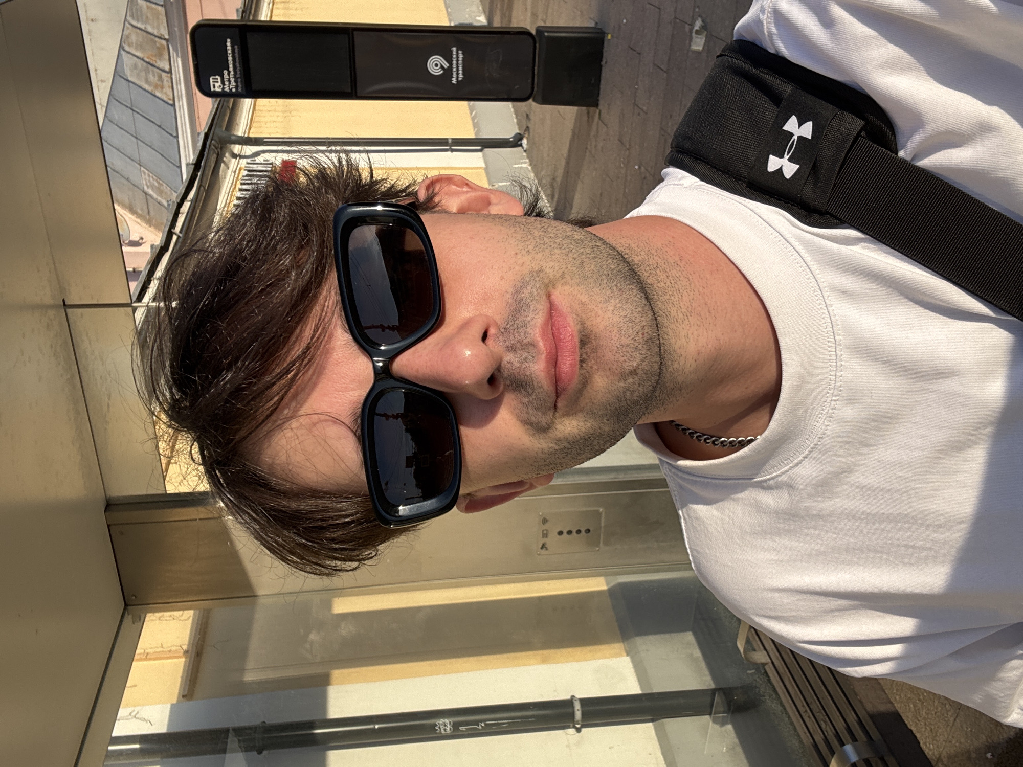 A man with dark hair and sunglasses stands in front of a bus stop in Moscow, Russia. He is wearing a white t-shirt and a black bag with the Under Armour logo is slung over his shoulder. Behind him, a yellow building and a black sign with the Moscow transport logo can be seen. The sun is setting, casting a warm glow on the scene.