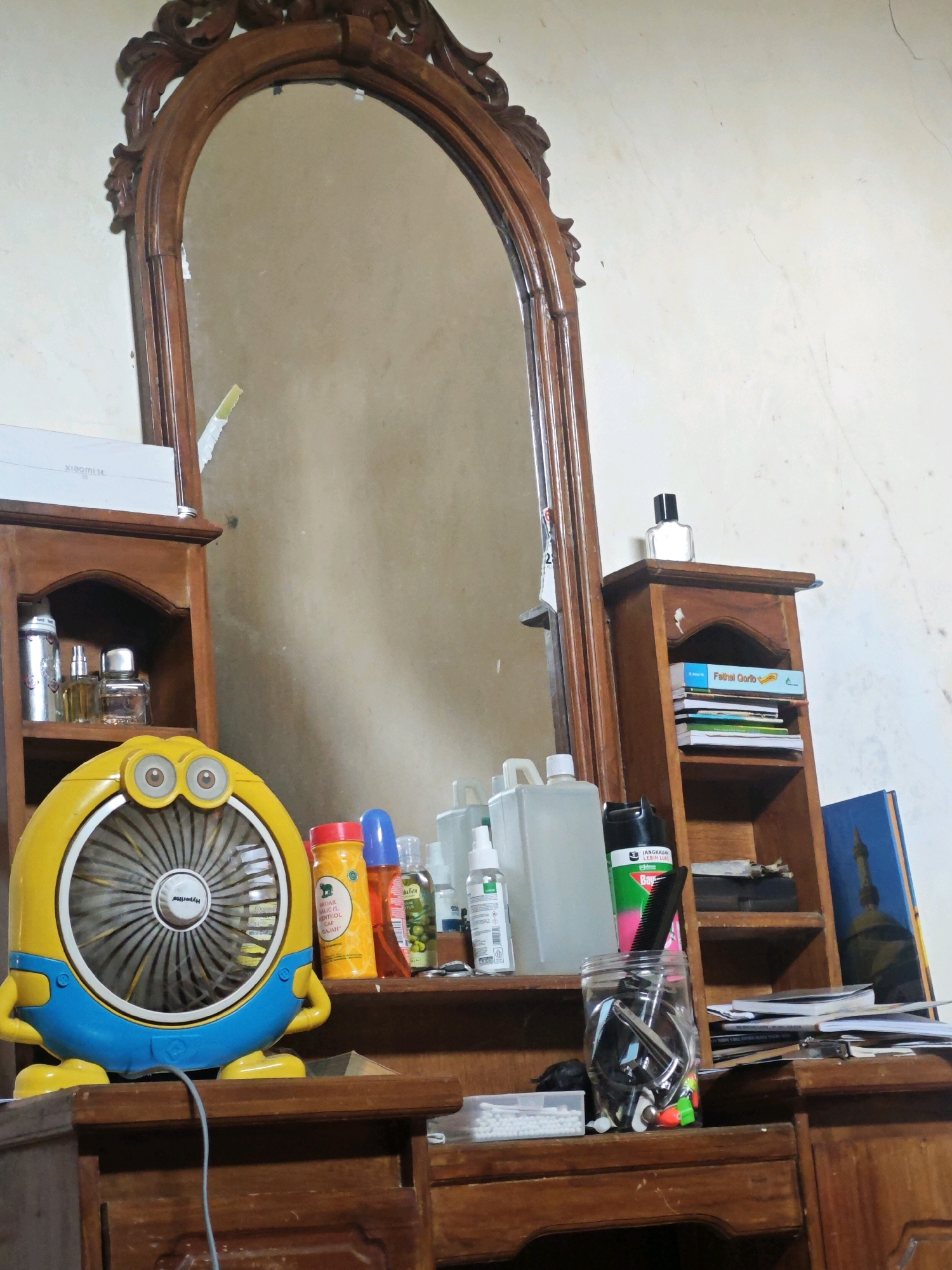 A dresser with a large, ornate wooden mirror and various items on its surface and shelves is shown. The dresser is made of dark wood and has several drawers. On the surface of the dresser, there is a yellow and blue fan shaped like a minion, a bottle of hair oil, a bottle of orange-colored liquid, and a blue bottle. Behind these items, on a shelf, are several bottles of perfume and a white box. To the right of the mirror, there is a wooden shelf unit with books, papers, a jar containing various items, and a can of spray. A framed picture is leaning against the shelf unit. The overall scene appears to be a personal dressing area, possibly in a bedroom or a vanity room. The lighting suggests it is daytime, with natural light coming from an unseen source.