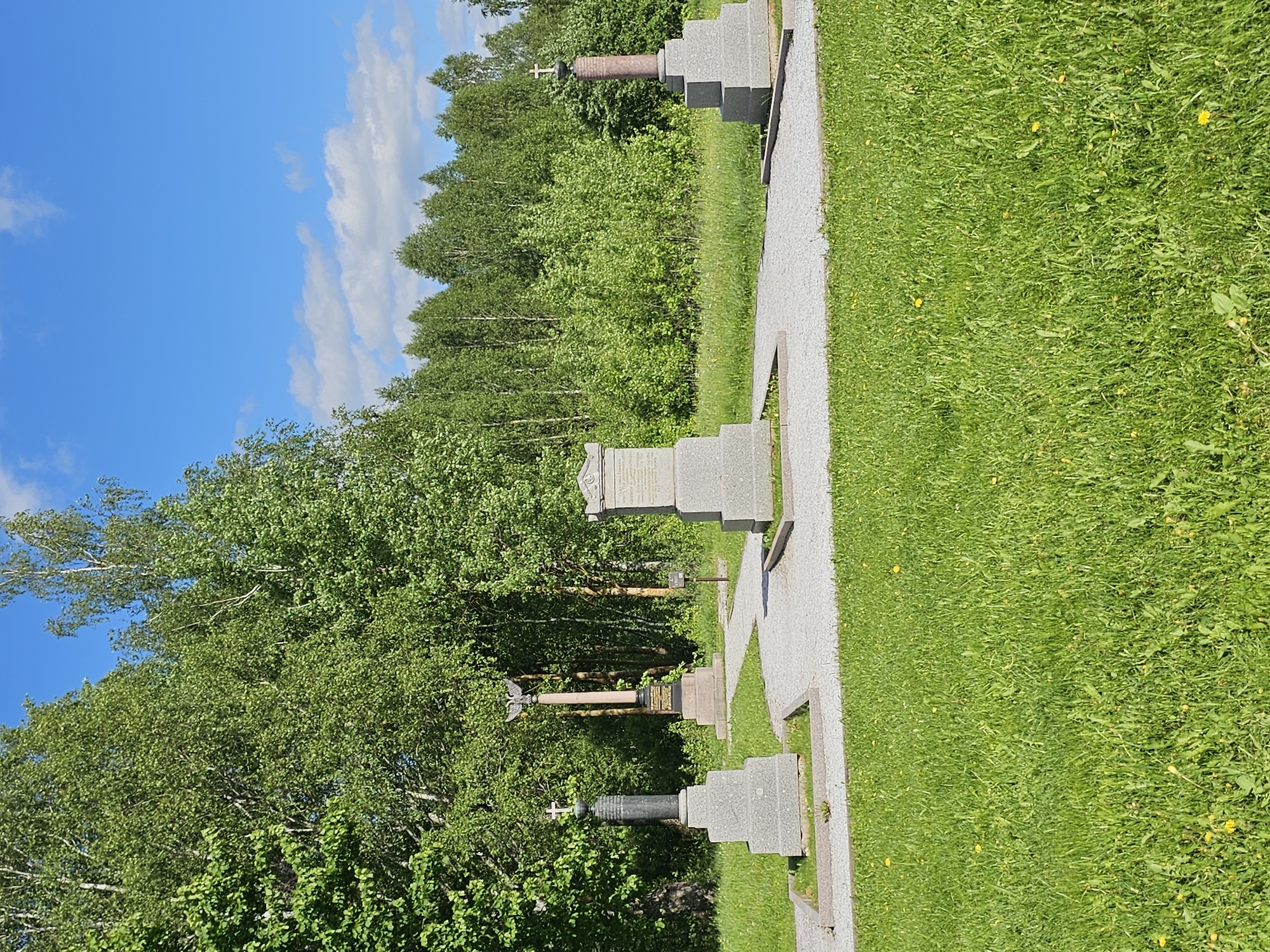 The image depicts a cemetery in Borodino, Russia. The cemetery contains various headstones and memorials arranged along a gray stone pathway bordered by green grass. Beyond the cemetery, lush green trees fill the background. The sky above is blue, dotted with scattered white clouds.
