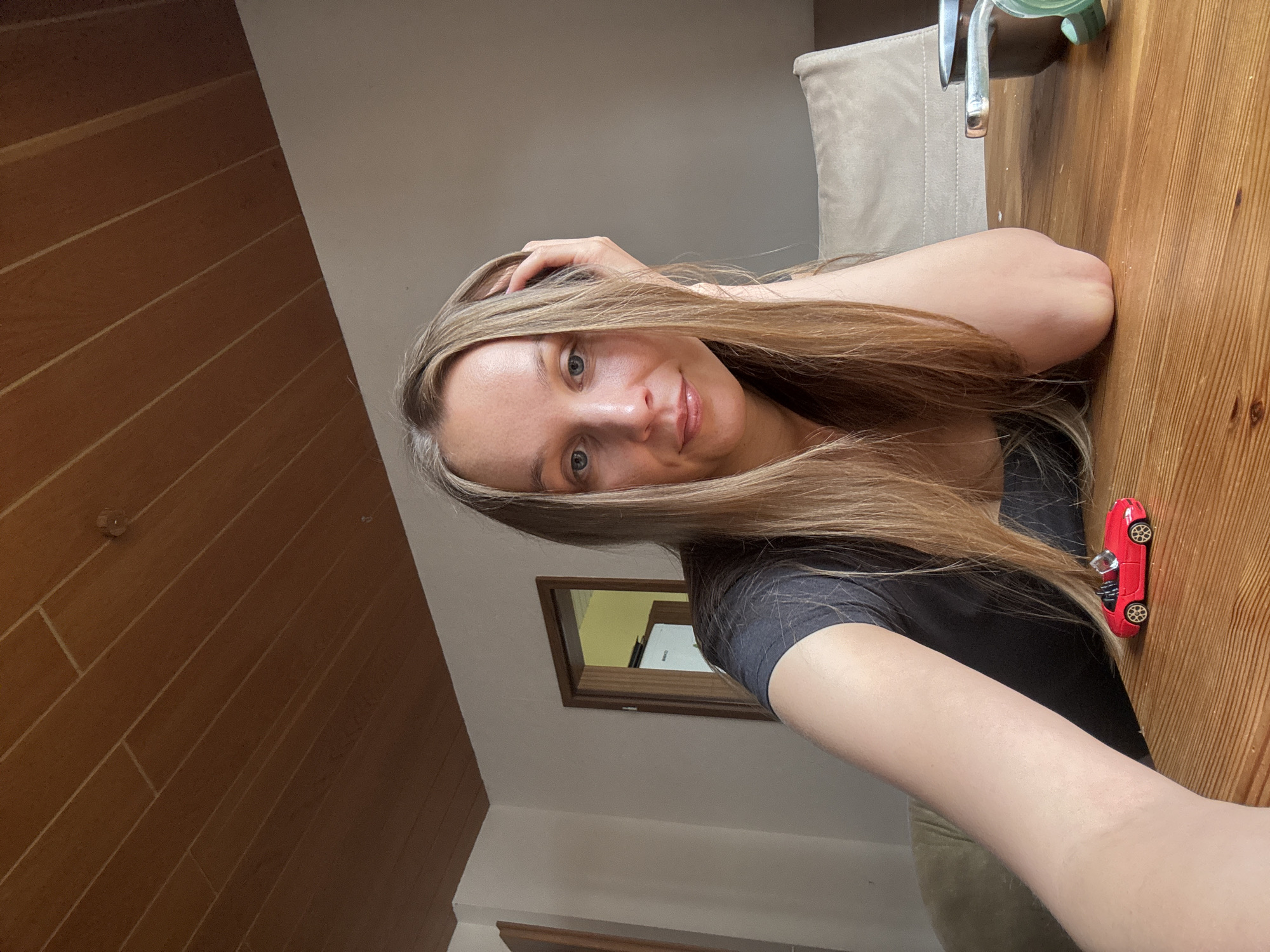 A medium shot, eye-level, selfie-style photograph captures a woman seated at a wooden table. The woman has long, blonde hair, fair skin, and blue eyes. She is wearing a dark gray t-shirt and resting her left arm on the table, supporting her head with her hand. To her right, on the table, is a small red toy car. The background features a wall, a doorway leading to another room, and a wooden paneled ceiling. The scene appears to be indoors, likely within a residence in Überherrn, Germany.