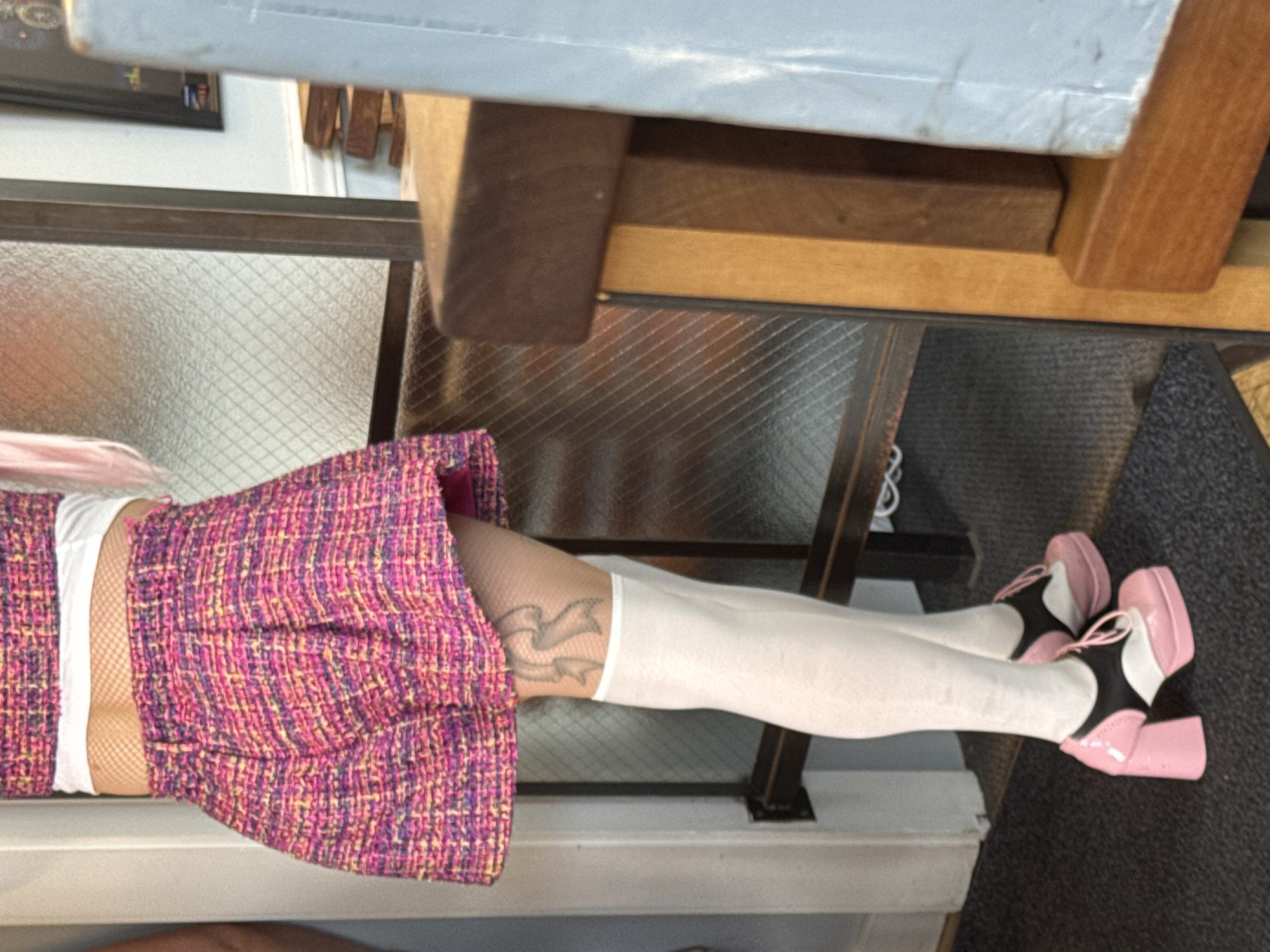 The image shows a person's legs and torso, viewed from the side and slightly from above. The person is wearing a textured pink and purple skirt, white stockings, and pink and white platform shoes with black accents. A tattoo of a ribbon design is visible on the left leg. The background appears to be indoors, with a metal railing and a textured surface that could be a carpet or floor. A wooden structure is visible behind the railing. The lighting is artificial and the image is oriented sideways. The overall impression is of a person posing, possibly for a fashion or personal photograph. No specific activity or event is clearly depicted. No text is visible in the image. The location context of Provincetown, USA, does not offer any specific cues within the visual itself.