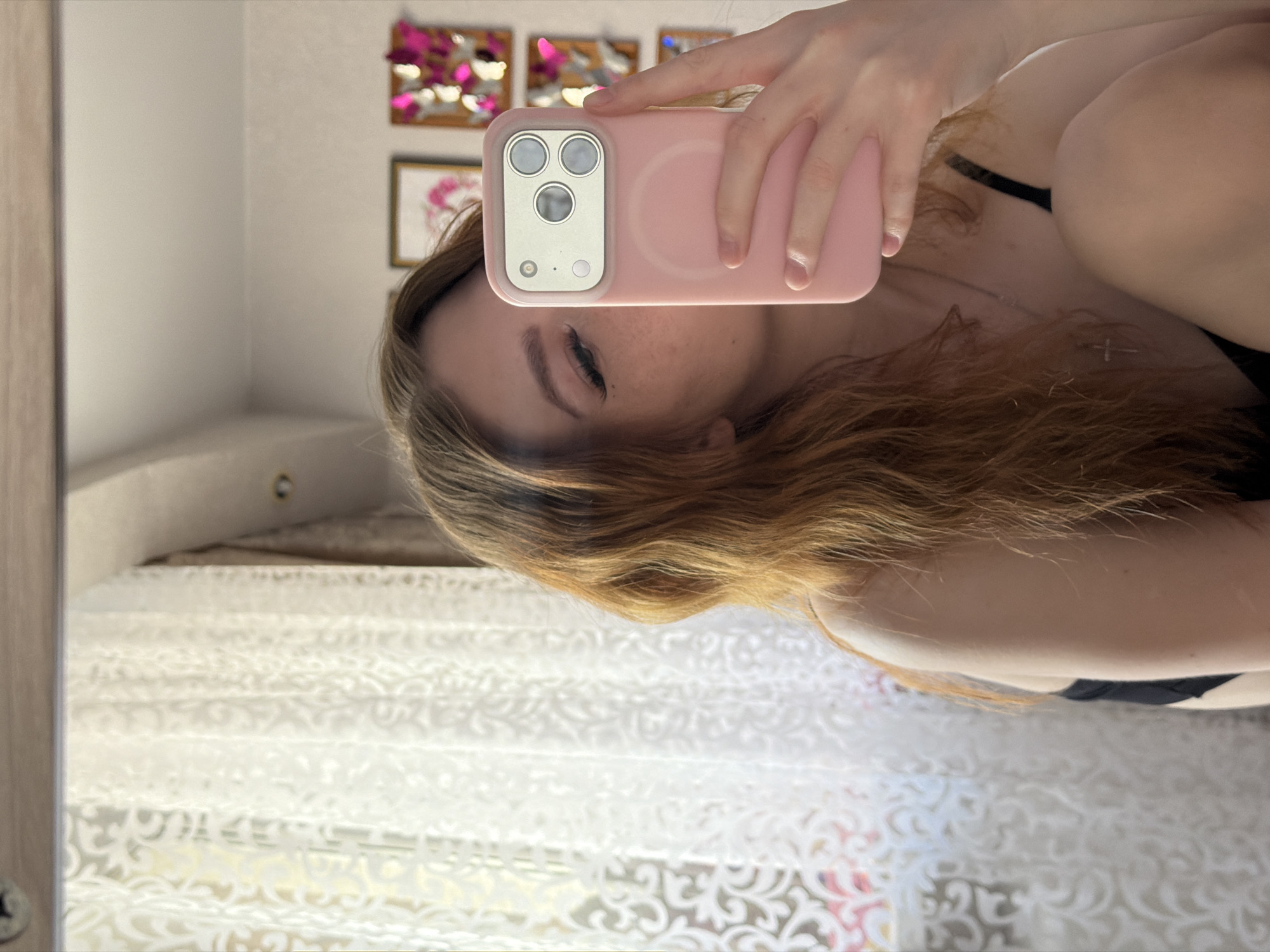 A woman is taking a selfie in a mirror. She is holding a pink phone in front of her face, obscuring most of it. Her hair is long and blonde. She is wearing a black top. The background shows a room with artwork on the wall and a patterned curtain. There is no information to suggest this photo was taken in the city of Noviy, Russia.