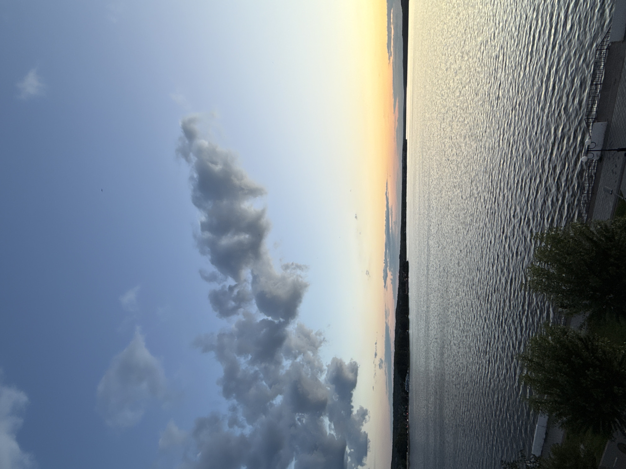 The image captures a serene landscape featuring a vast body of water, likely a lake or reservoir, under a sprawling sky during late afternoon or early evening. The horizon line cleanly divides the scene into sky and water.
The sky, occupying the upper portion of the frame, transitions from a clear, light blue on the left to warmer, soft yellow and orange tones near the horizon on the right, suggesting the approach of sunset or the aftermath of sunrise. Various cumulus clouds, ranging from soft white to darker grey, are scattered across the sky, with one crop revealing a distinct, tear-drop shaped cloud. A tiny, distant speck, possibly a bird, is faintly visible in the expanse of the blue sky.
The lower half of the image is filled by the calm surface of the water, which displays subtle ripples and a shimmering texture under the diffused light. The water appears as a dark, muted blue-grey, reflecting the ambient light. Along the distant horizon, a dark silhouette of land, possibly a far shoreline with hills or a town, is visible, with faint artificial lights discernible against the fading light.
In the foreground, on the bottom right of the main image, a section of a developed waterfront area is partially visible. This includes some dark green foliage, likely bushes or trees, and a portion of a lamppost equipped with two lights. The structure of the embankment or promenade appears to be made of concrete or stone.
The scene, devoid of human activity, evokes a sense of peacefulness and natural beauty. The time of day is clearly indicated by the color gradient in the sky. The setting strongly suggests a large body of water, consistent with the Votkinsk Reservoir in Russia. No visible text is present.