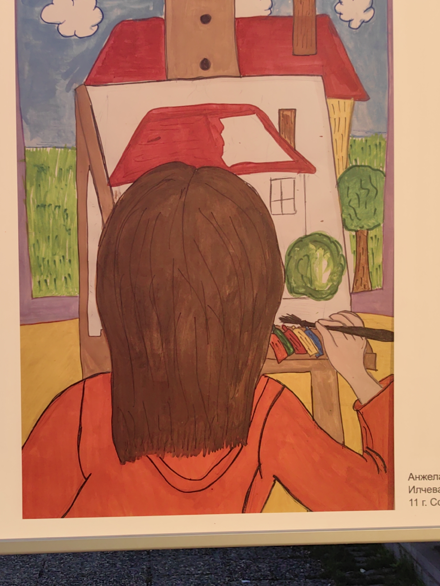 The image shows a framed artwork, likely a child's painting, depicting a person with brown hair, wearing an orange shirt, painting a picture of a house. The painting is on an easel. The house in the painting has a red roof, a window, and a chimney. Behind the house, there is a blue sky with clouds, grass, and trees.
The artwork is displayed in a location where the artist's name and age "Анжела Илчева, 11 г. София" are visible, indicating the artist is Angela Ilcheva, an 11-year-old from Sofia, Bulgaria.