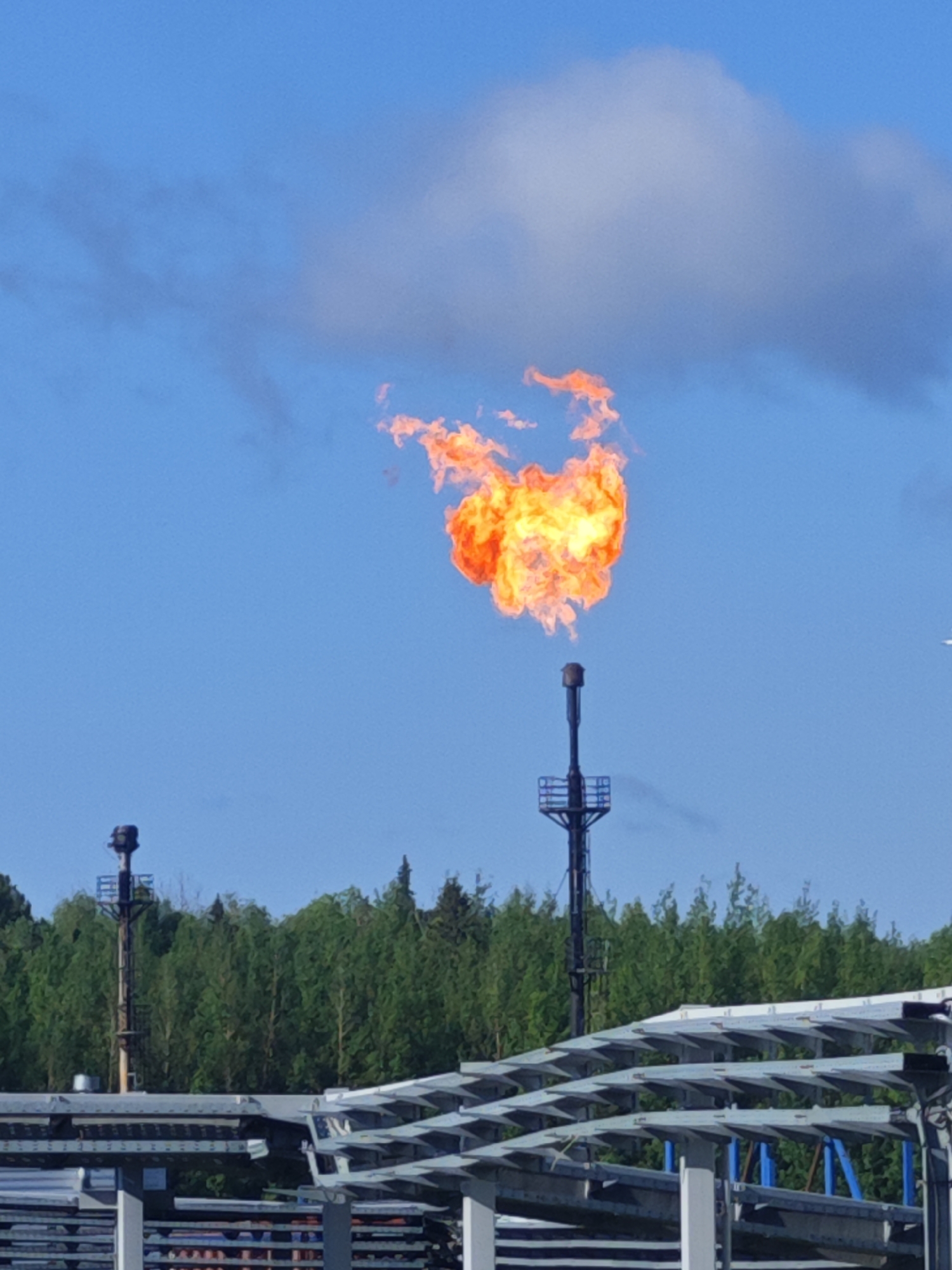 A large, bright orange flame flares from a tall, narrow stack, reaching upwards into a clear blue sky. The flame is the dominant feature, with swirling patterns of yellow and red. In the background, a dense line of green trees forms a natural horizon. In the foreground, there are several levels of grey, angular structures, possibly part of an industrial complex. To the left, another, shorter stack is visible amidst the trees. The overall impression is one of industrial activity against a natural backdrop, with the flame symbolizing energy production or venting. The location is unknown, but the presence of industrial structures and a forest suggests a rural or industrial outskirts setting.