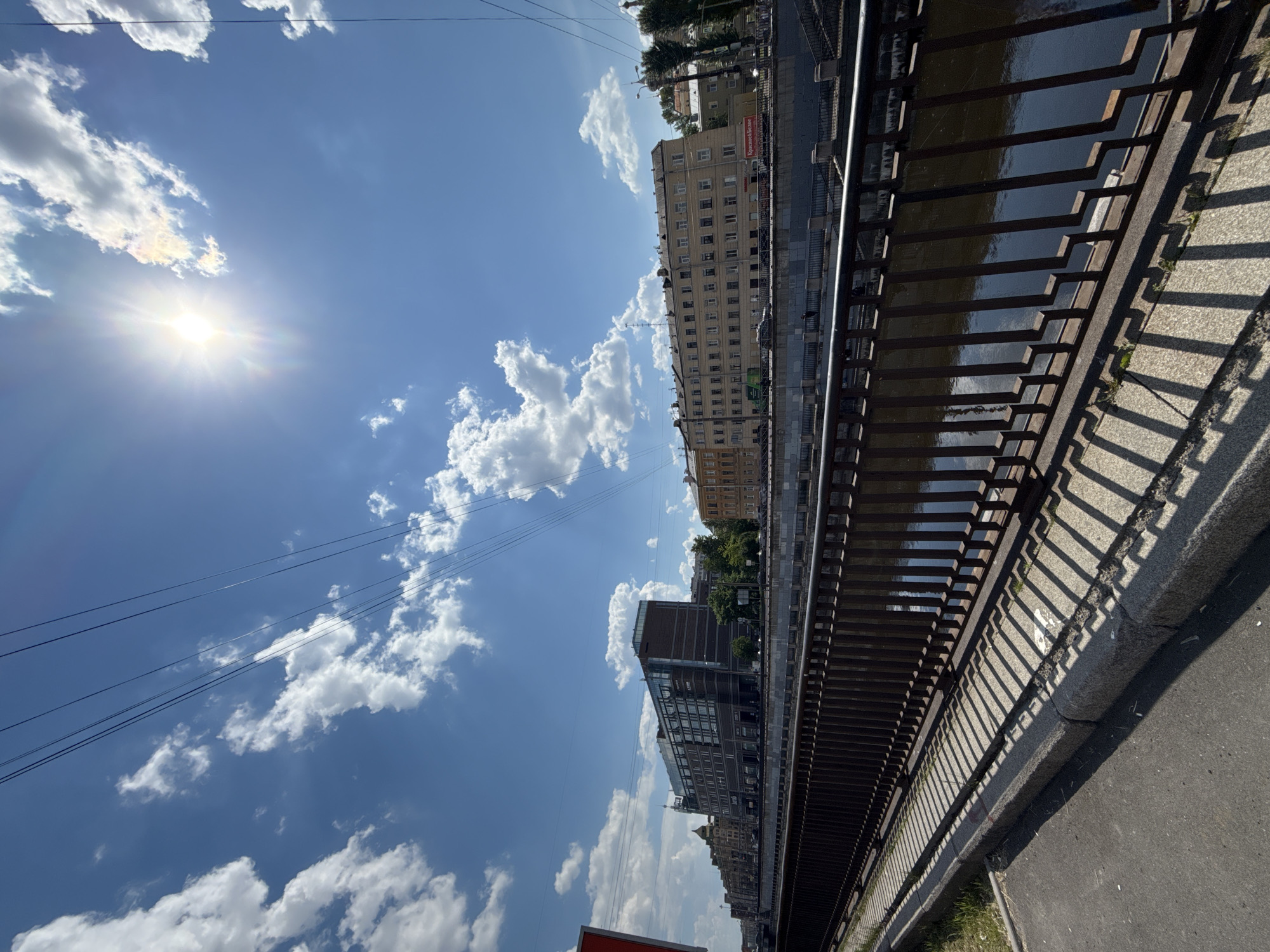 The image depicts an urban landscape under a bright, sunny sky. The perspective is from a waterside promenade or embankment, looking across a canal or river toward a row of buildings.
In the foreground on the right, a dark metal railing runs horizontally, separating the viewer from the water. This railing casts strong, angular shadows onto the adjacent concrete pavement and a section of asphalt road. The water in the canal or river appears dark and reflective, visible through the gaps in the railing.
Across the waterway, a series of multi-story buildings forms the opposite bank. These buildings vary in architectural style; some are older, light-colored structures with numerous windows, while others appear more modern with darker facades and larger windows. On one of the more prominent, lighter buildings, a red sign with white text is clearly visible, reading "Красное & Белое".
The sky dominates the upper portion of the image, a vivid blue with scattered, fluffy white cumulus clouds. The sun is a brilliant burst of light in the upper left quadrant, creating sharp highlights and deep shadows throughout the scene. Several thin power lines or overhead cables stretch across the sky in the middle ground.
The time of day is clearly daytime, likely midday or early afternoon, given the intensity of the sun and the distinct shadows. The weather is bright and clear. No people are visible, focusing attention on the architectural and environmental elements. The visible text "Красное & Белое" suggests the location is in Russia.