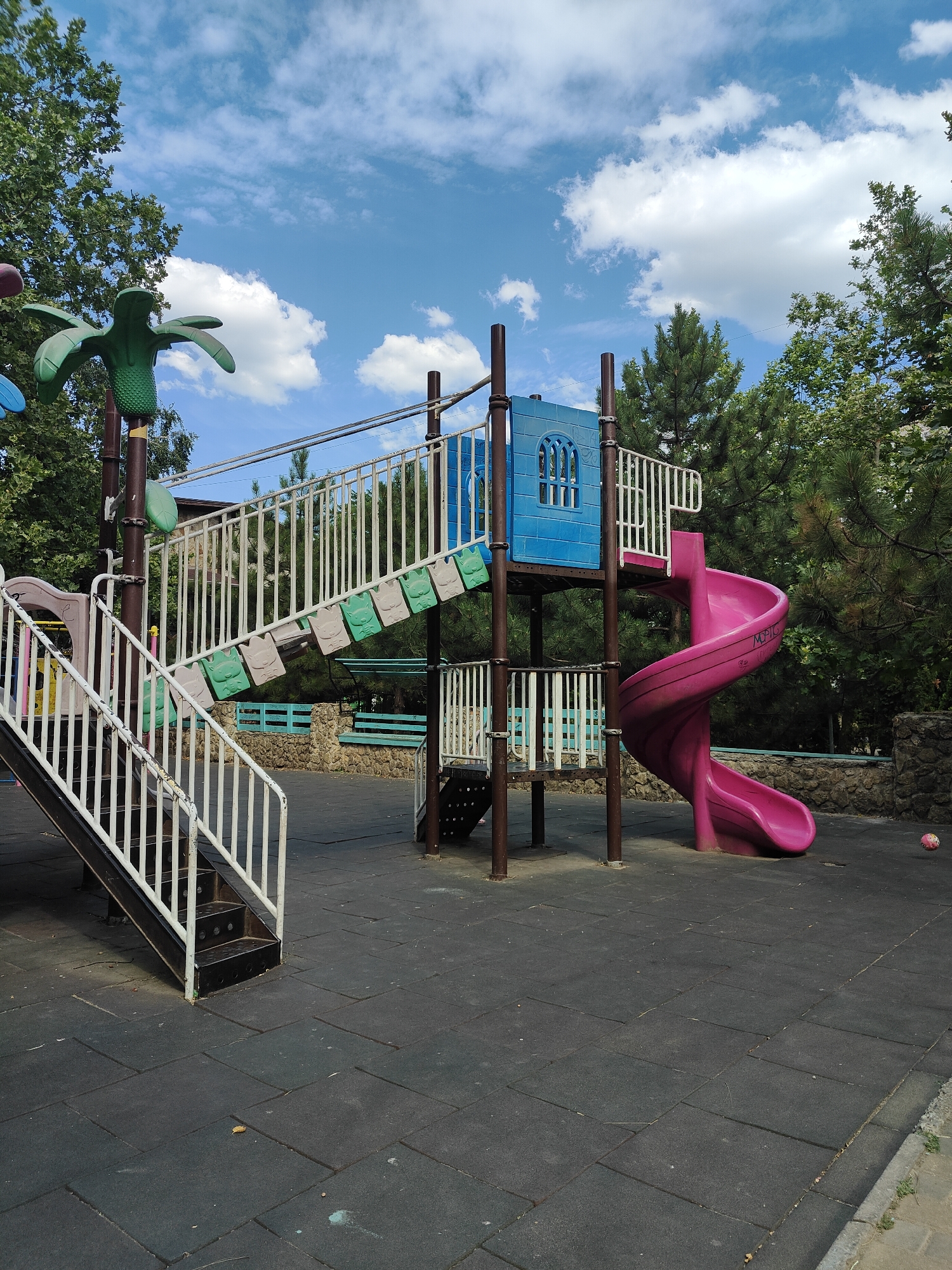 The image shows a playground in Chornomorsk, Ukraine, under a blue sky with scattered white clouds. The playground equipment includes a blue structure with windows, a spiral pink slide, and stairs with white railings. Green palm-tree-shaped decorations are visible on the left side of the structure. The ground is covered with black rubber tiles. Trees surround the playground, and a stone wall is visible in the background. A pink ball is on the ground to the right of the slide. There are no people visible in the image.