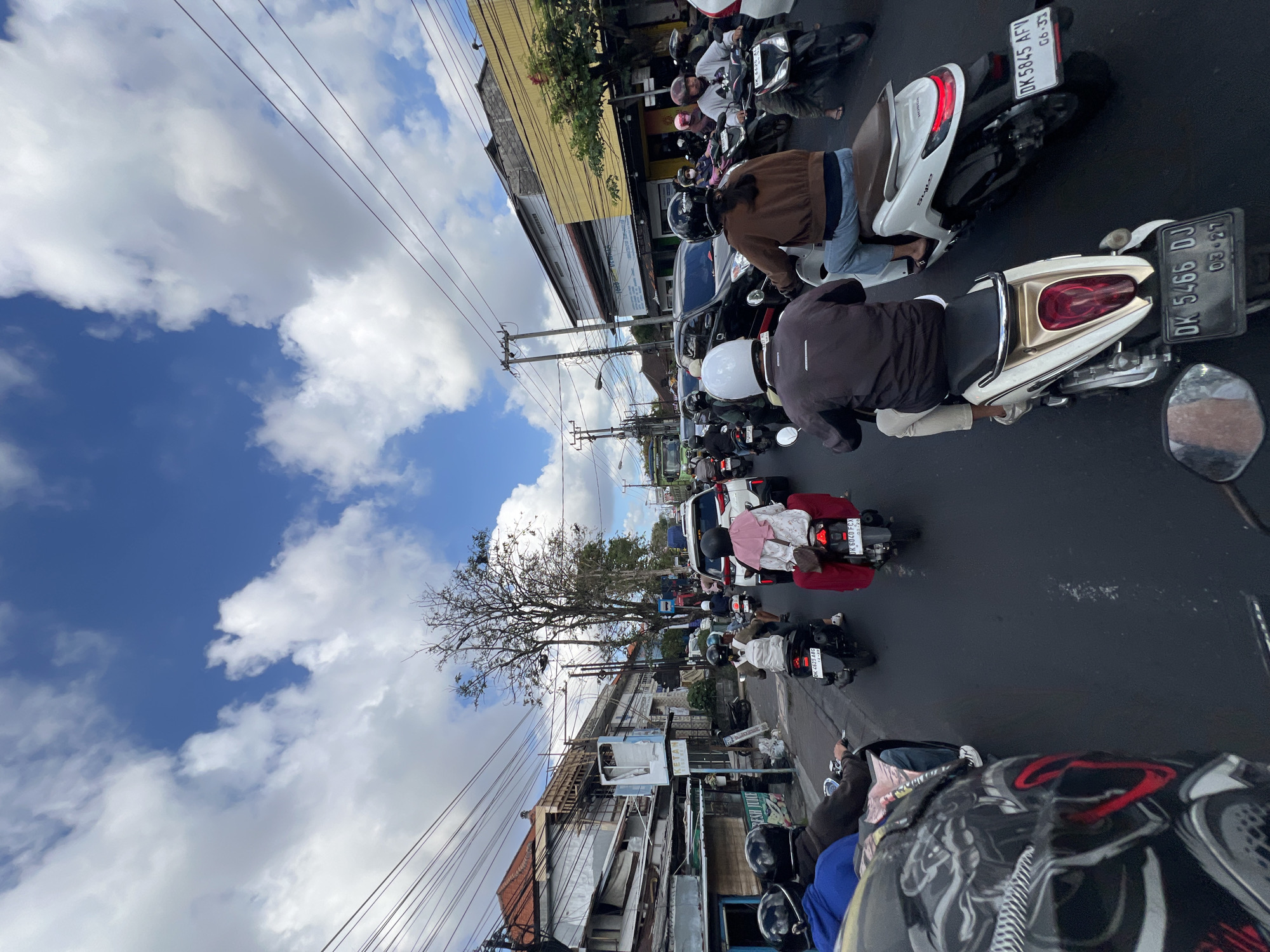 The image shows a congested street scene in Denpasar, Indonesia. Multiple motorbikes and some cars are moving slowly through traffic. The sky is a vibrant blue with scattered white clouds. Buildings line the sides of the street, with power lines and utility poles overhead. Many people on the motorbikes are wearing helmets. One motorbike in the foreground has a license plate that reads "DK 5466 DJ". Another motorbike in the middle ground has a license plate reading "DK 6040 FCX". The overall impression is of a busy, urban environment with a lot of vehicle and pedestrian activity.
