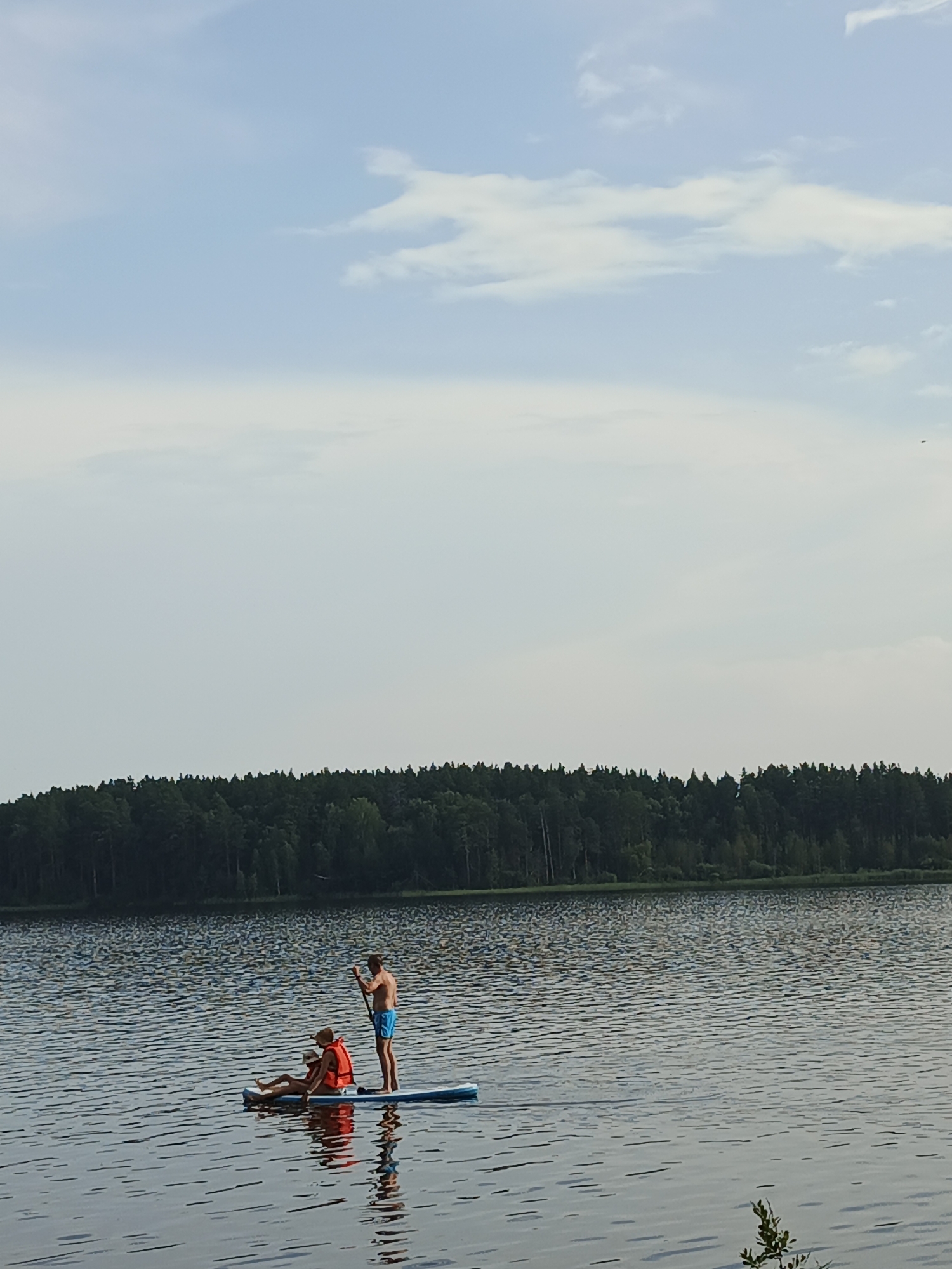 In the city of Sarafanovo, Russia, a serene lake scene unfolds. The sky above is a pale blue, with scattered white clouds. In the middle ground, a dense forest lines the far shore. The foreground is dominated by the calm, rippling surface of the lake.
On the lake, a blue and white paddleboard is occupied by two individuals. A man, wearing blue swim trunks, stands at the rear of the paddleboard, holding a paddle. He appears to be propelling the board forward. In front of him, a person wearing a bright orange life vest and a hat is seated on the paddleboard. Their legs are extended, and they are looking towards the water. The reflection of the paddleboard and its occupants is visible on the lake's surface. The overall atmosphere suggests a peaceful recreational activity on a calm day.