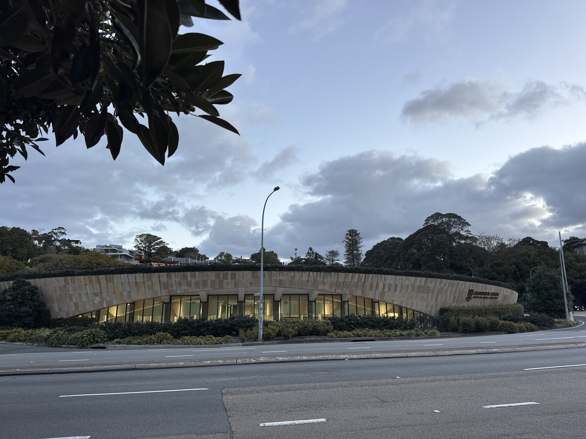 The image shows a curved, modern building with a beige stone facade and large glass windows. The building is set against a backdrop of trees and a cloudy sky. The foreground features a road with lane markings. The building appears to be illuminated from within. A street lamp is visible to the left of the building. The building has a sign that reads "CRANBROOK SCHOOL" and a crest. The overall scene is a street-level view of the school building.