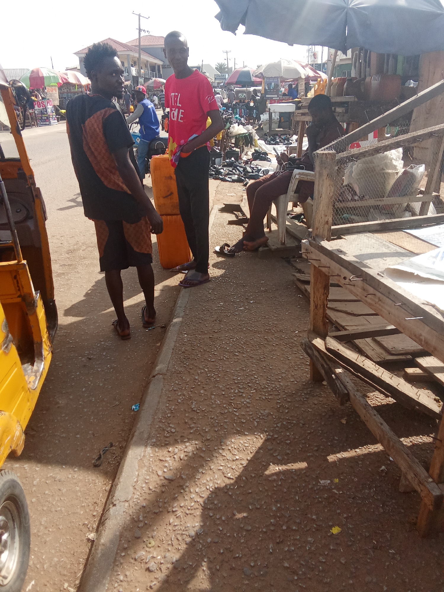 Two men stand near a yellow auto-rickshaw in what appears to be a street market in Kubwa, Nigeria. The man on the left wears a black and orange patterned t-shirt and shorts, with dark sandals. He is looking away from the camera, with his right arm bent at the elbow. The man on the right wears a bright red t-shirt with the word "ITALIE" printed in white, and black pants. He is facing the camera and holding a white object in his left hand. They are standing on a dirt and gravel surface next to a curb. Behind them, two large orange containers are visible, along with various market stalls, umbrellas, and other people engaged in market activities. The overall scene suggests a busy outdoor market environment.