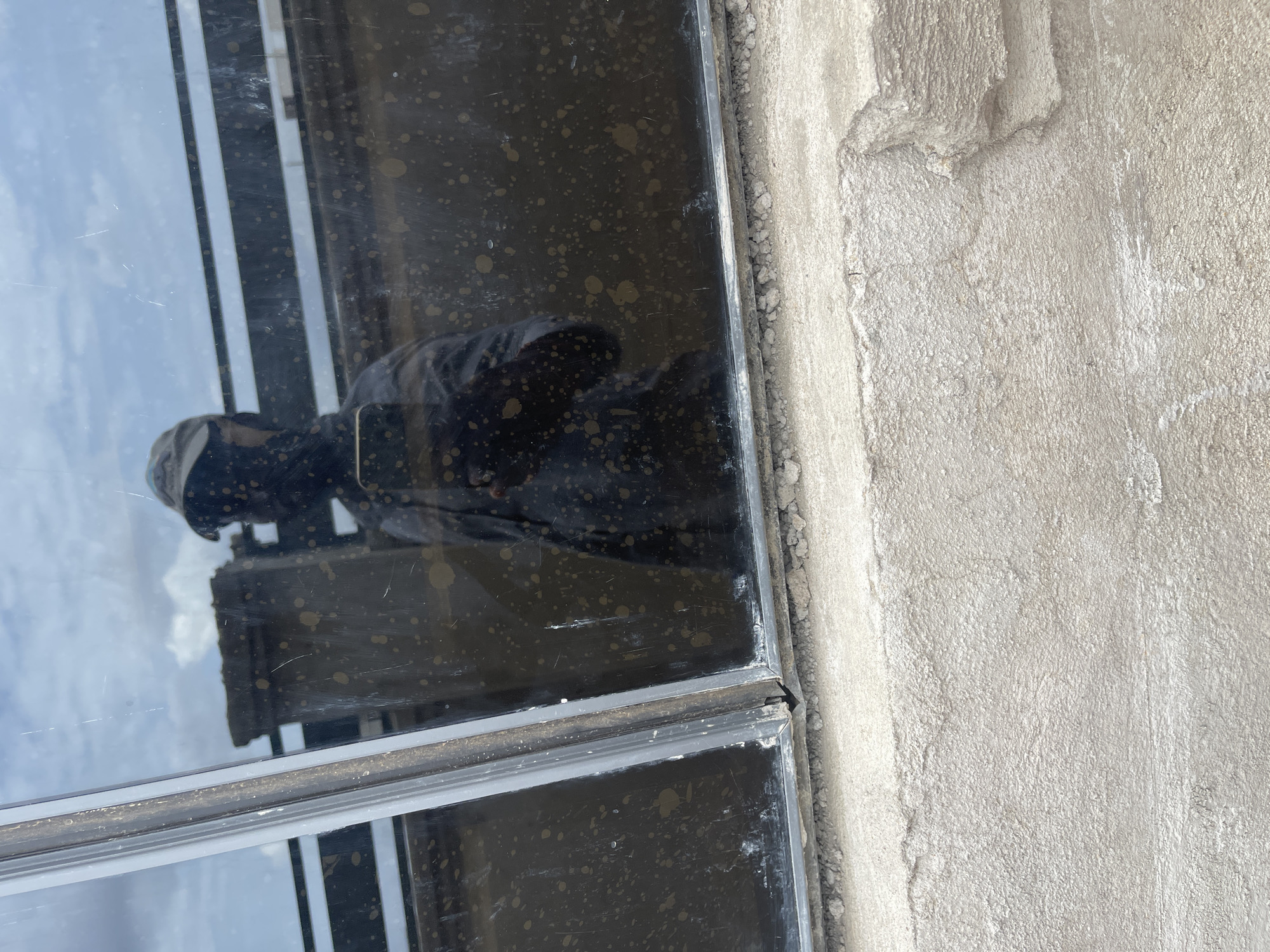 A close-up shot shows a dirty window frame embedded in a rough, light-grey wall. The window glass reflects a person holding a phone, presumably taking the picture. The location, Angwan Sayawa, Nigeria, is not directly discernible from the visual data.