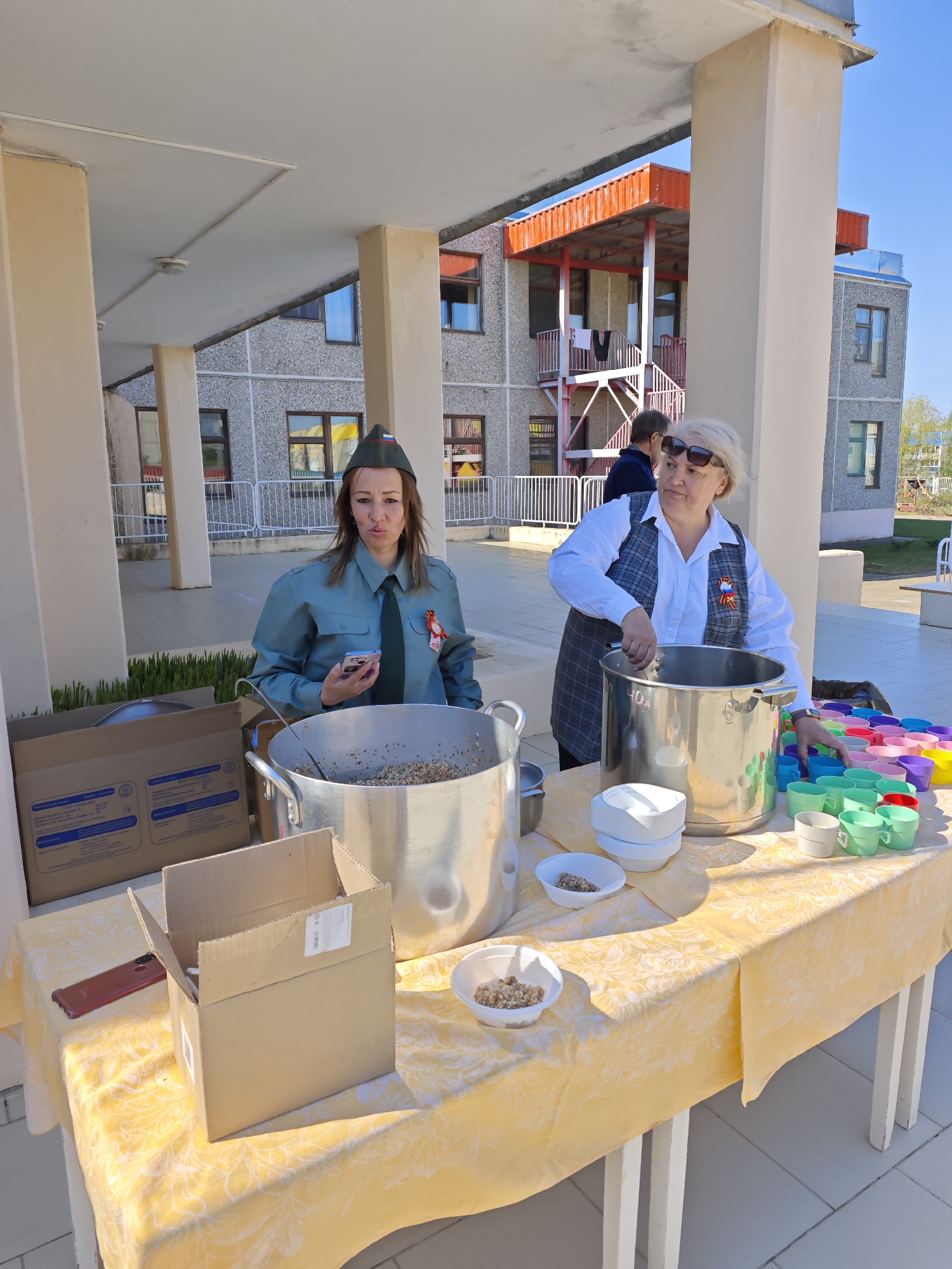 The image depicts two women standing behind a table outdoors, seemingly serving food. The location is the city of Markovskii, Russia, as suggested by the uniform and other items on the table.
The woman on the left wears a green military-style uniform, including a garrison cap, and a ribbon pinned to her chest. She is holding a cell phone in her left hand and looking toward the camera. Next to her is a large metal pot on the table. A cardboard box is also visible to her left.
The woman on the right is wearing sunglasses and a white long-sleeved shirt under a gray patterned vest. She has a ribbon pinned to her chest. Her right hand is inside a large metal pot in front of her. A stack of white bowls and a collection of colorful cups are on the table next to the pot.
The table is covered with a yellow patterned tablecloth. There are bowls of food on the table and a partly disassembled cardboard box on the left.
The background features a building with beige columns and a red-trimmed second story, under a clear blue sky.