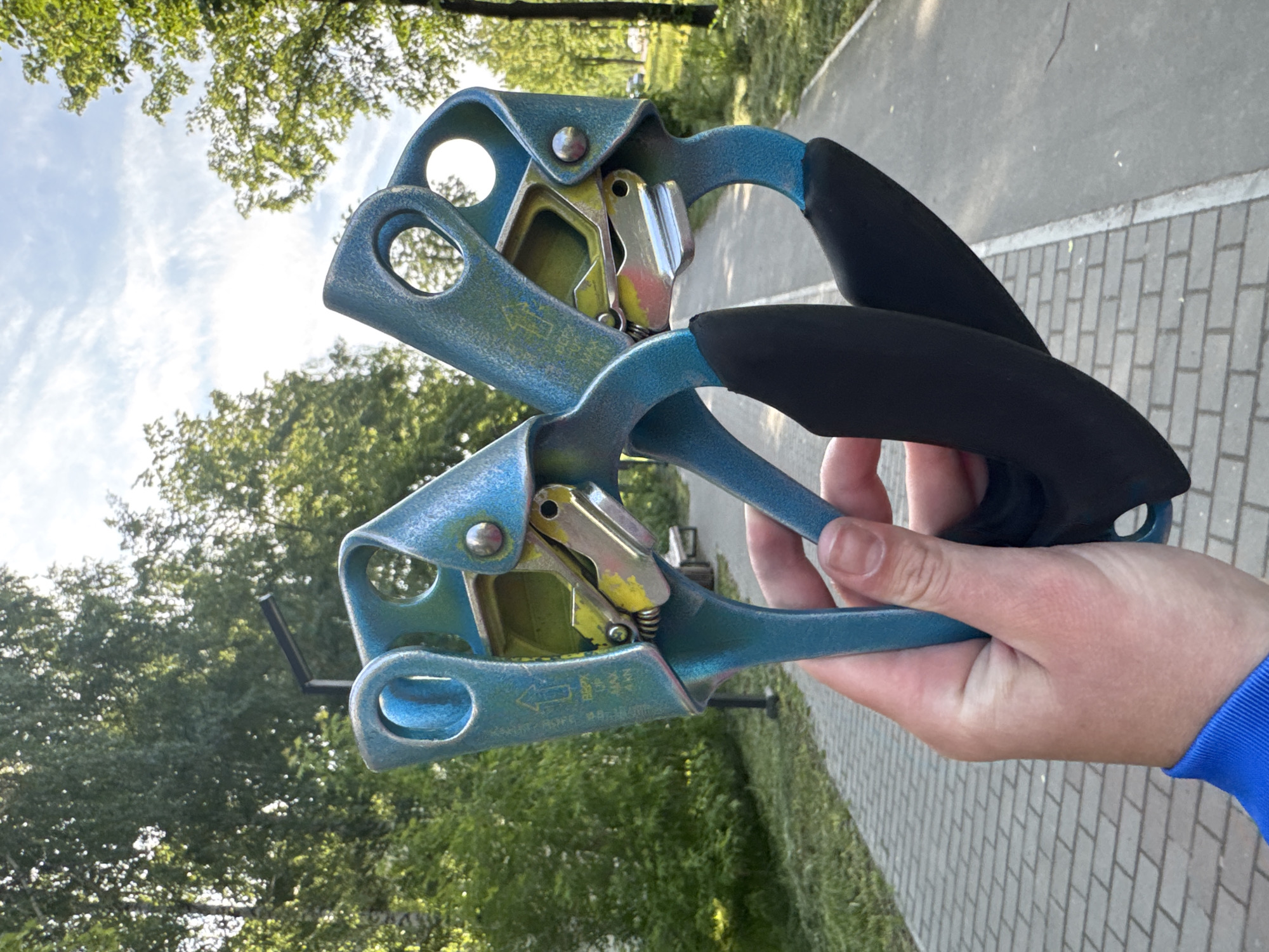 The image, captured in Chaikovsky, Russia, features a person's right hand holding two blue and yellow ascenders, which are climbing or rope access devices. The ascenders, appearing to be the same model, consist of blue metal bodies with black rubberized grips. Their internal mechanisms, including metallic cams and springs, are visible, with yellow paint on the internal parts showing signs of wear from use.
The background reveals an outdoor setting under bright daylight, likely during the morning or late afternoon. Lush green trees with dense foliage are prominent on the left and in the upper background, suggesting a park or wooded area. To the right and in the lower foreground, a paved path is visible, comprising both smooth gray sections and parts with a lighter, rectangular brick-like pattern. The sky above is partly cloudy and bright.
On the lower ascender, an arrow pointing left is clearly visible. Below it, partially legible text includes "ROPE" and "Ø 8-13 MM," indicating the recommended rope diameter for the device. The image primarily focuses on the equipment, with the hand serving to present the ascenders in an outdoor, recreational or operational context.