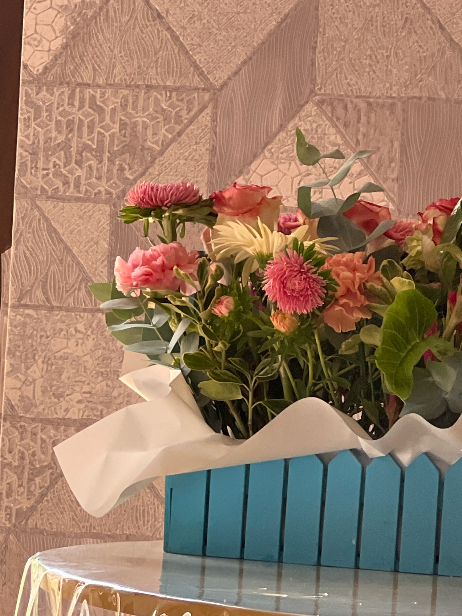 The image shows a close-up of a bouquet of flowers in a blue wooden planter. The flowers are a mix of pink and orange hues, with some green foliage visible. The planter is placed on a table covered with a clear plastic tablecloth. In the background, there is a textured wallpaper with a geometric pattern. The overall impression is one of a still life, with the focus on the floral arrangement. The scene appears to be indoors, with the lighting suggesting it might be daytime. There is no indication of Sokhumi, Abkhazia, or any specific location within the image.