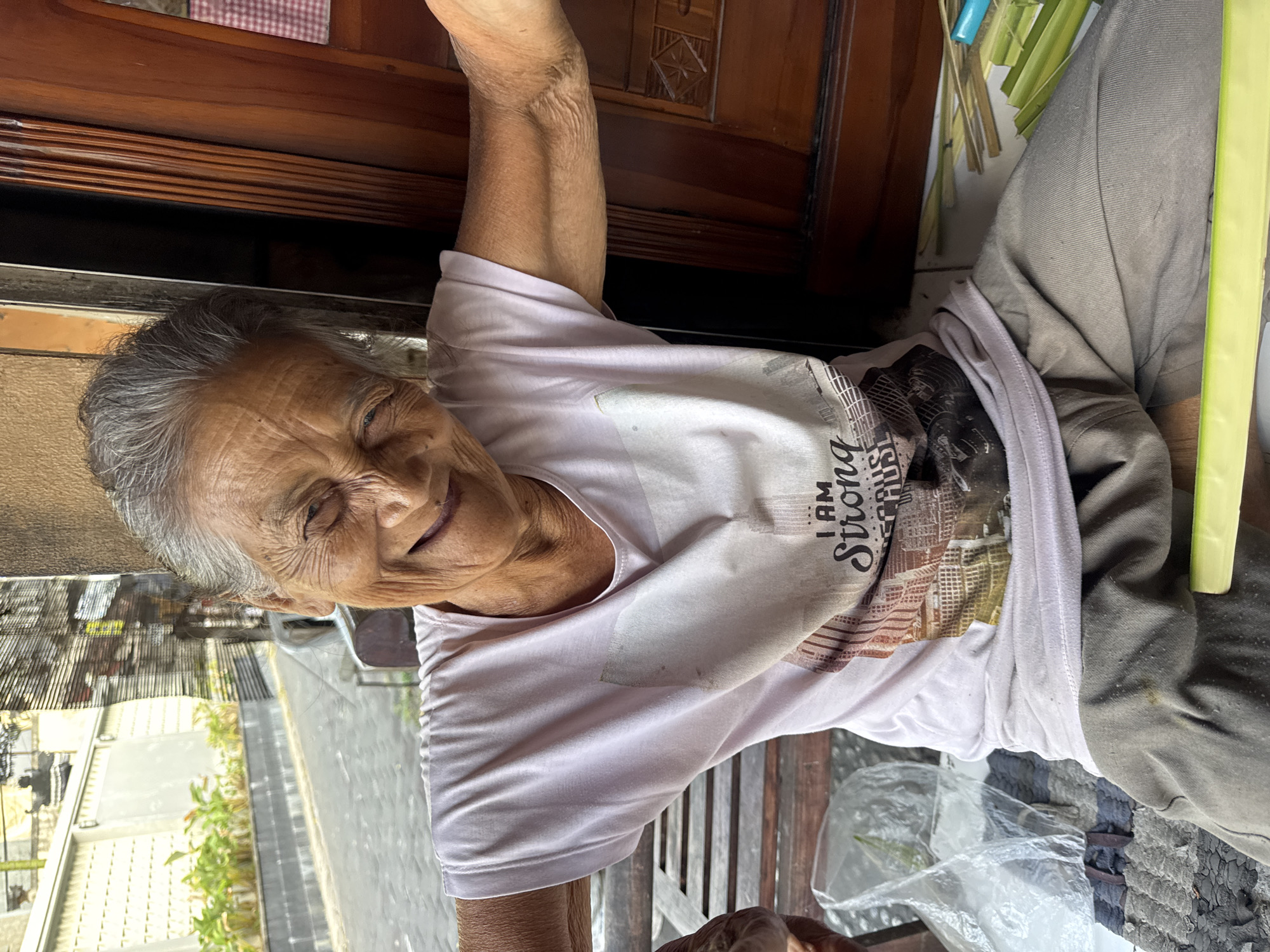 An elderly woman with gray hair and wrinkled skin is seated outdoors, smiling broadly with her eyes closed. She is wearing a light pink t-shirt with a graphic print that says "I AM STRONG" and loose-fitting gray pants. Her arms are extended to the sides. She is sitting on a mat with a plastic bag next to her. In front of her, a bundle of green leaves and sticks is visible. The background shows a street scene with a white wall, some plants, and a wooden door. This image appears to be taken in Kuta, Indonesia, likely a residential area given the setting.