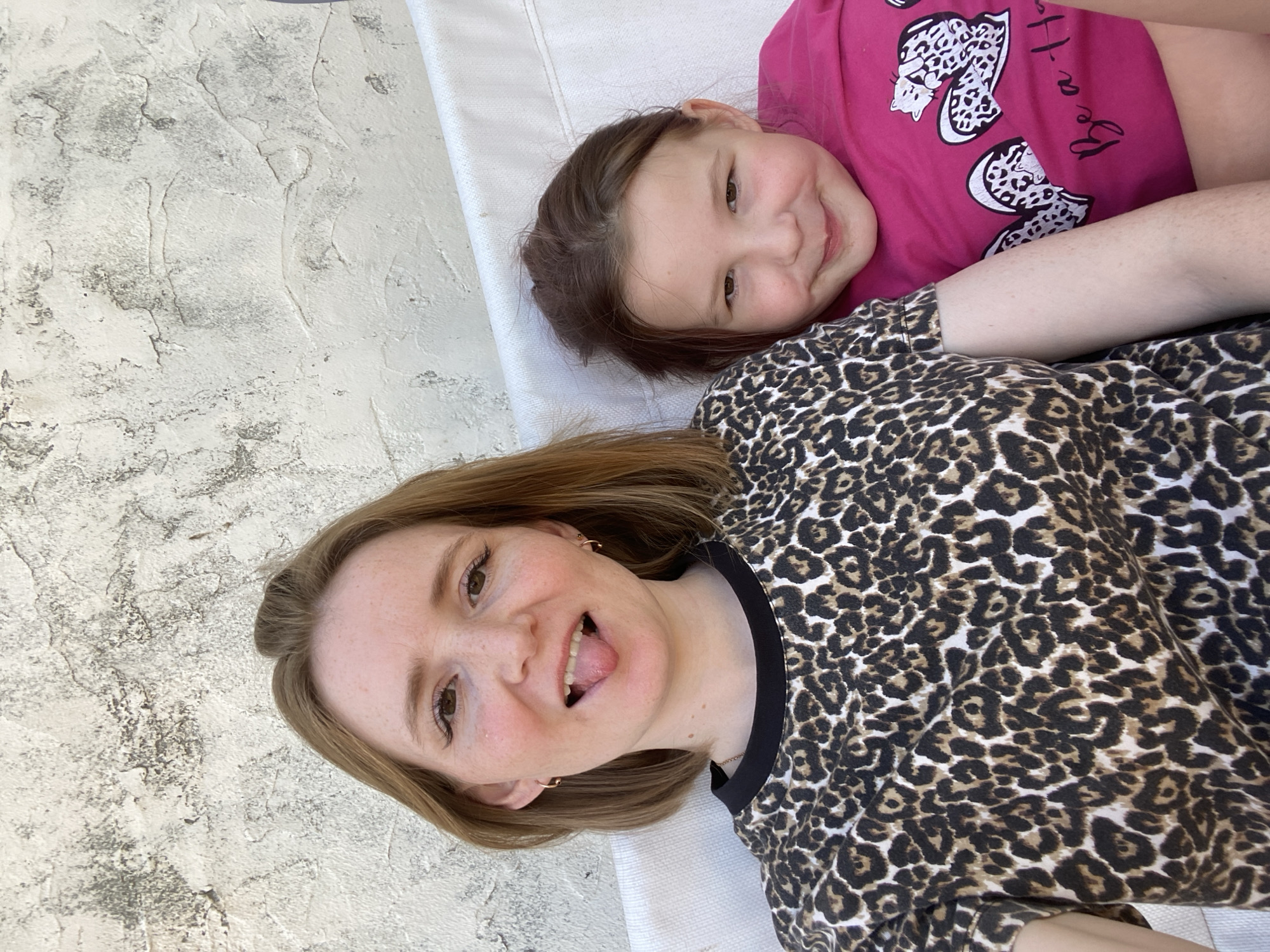 An outdoor, medium shot shows two females, an adult and a child, seated side-by-side in front of a textured, off-white wall. The adult, on the left, has reddish-brown hair and is wearing a leopard-print shirt with a black neckline. She is sticking her tongue out playfully. The child, on the right, has brown hair and is wearing a bright pink t-shirt with cat and heart designs and the text "Be a Ha". The child is smiling at the camera. The two subjects appear to be on a bench or sofa, suggested by a white fabric in the foreground. The lighting indicates it is daytime, with sunlight casting shadows and highlighting the textured wall. The location is not discernible from the image.