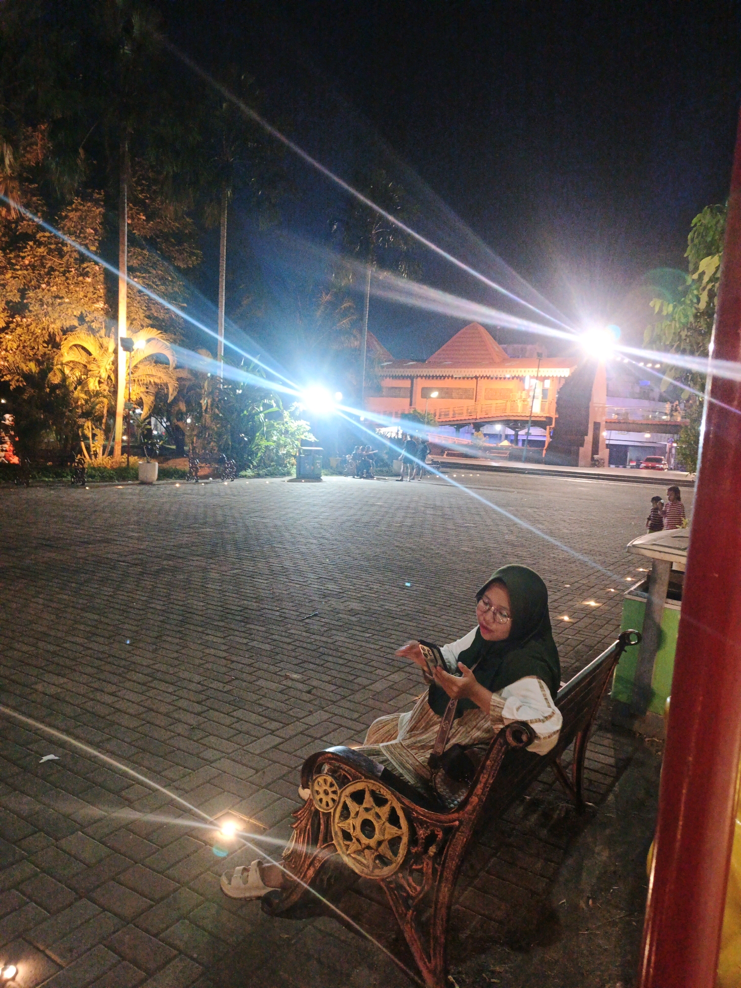 A young woman wearing a dark green hijab and glasses is seated on an ornate metal bench at night. She is looking down at her smartphone, which she holds in her right hand, and appears to be typing or browsing. She is wearing a white top with decorative sleeves and a patterned skirt. The bench has intricate metalwork on its arms and legs, including starburst designs.
The setting is an outdoor paved area at night, likely a public space in Mojokerto, Indonesia, given the architectural style of the building in the background, which resembles a traditional Indonesian structure with a tiered roof. The area is illuminated by bright lights, causing lens flares and strong highlights. Lush tropical foliage, including palm trees, is visible in the background, further suggesting a warm climate. The ground is paved with interlocking bricks. Other people are visible in the background, some standing and others seated, suggesting a social gathering or public event. A red cylindrical object is prominent in the foreground on the right side of the image.