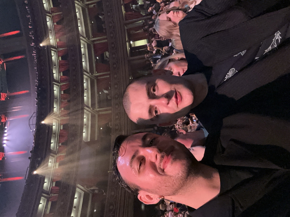 Two men take a selfie in what appears to be an ornate theater. One man has a shaved head, the other has dark hair slicked back. Both are wearing black shirts. The theater is full of people in the background. Stage lighting illuminates the scene, and rows of seats and architectural details such as curtains in the balconies are visible.