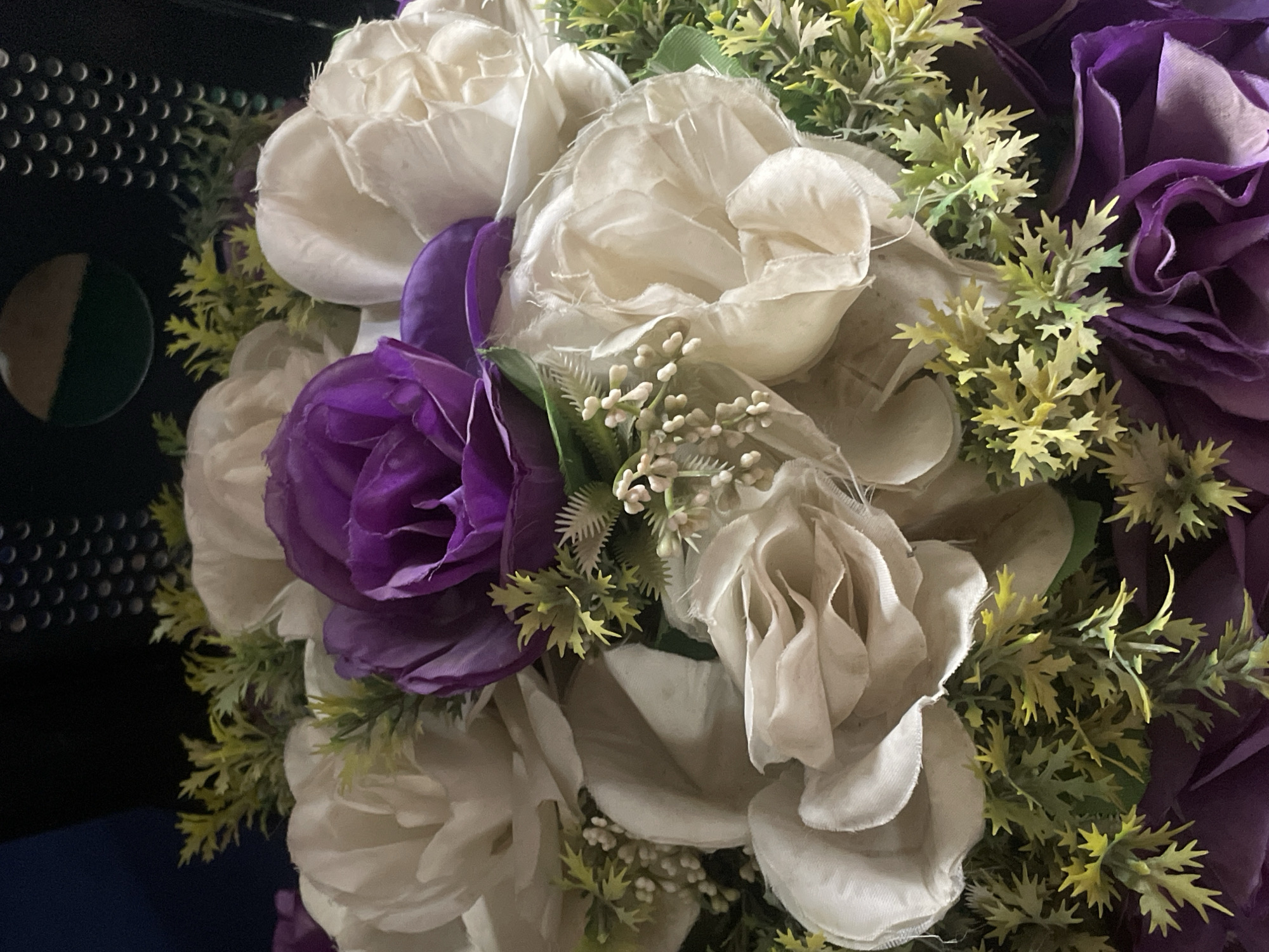 The image presents a close-up view of an arrangement of artificial flowers. Predominantly, there are white and purple rose-like blooms. Greenery, including small leafy stems, is interspersed among the flowers. The background is blurred, showing part of a perforated black surface and a dark blue or black object. The photo shows no people. It is impossible to ascertain if this photo was taken in the city of Jos, Nigeria, or anywhere else.