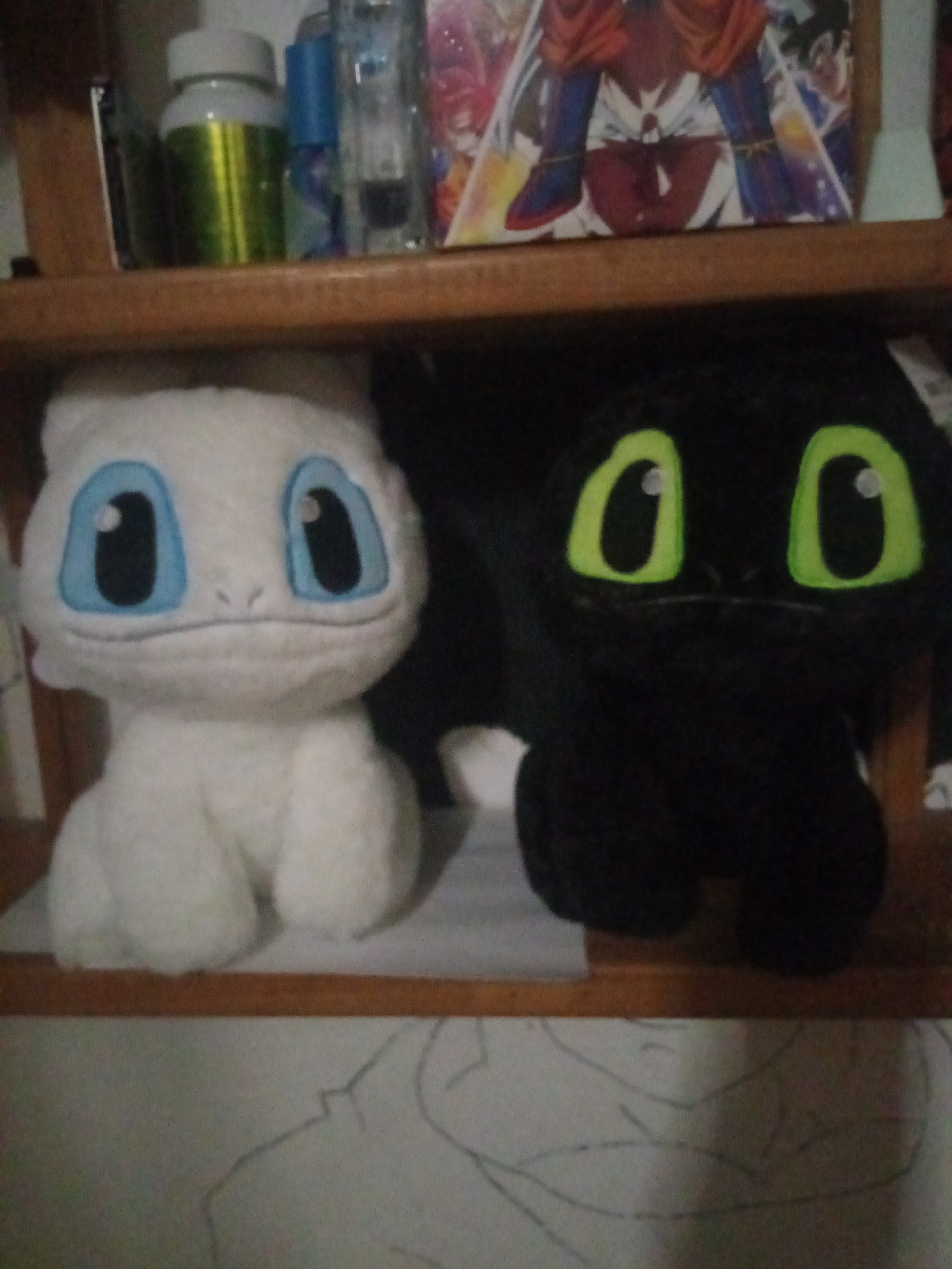 The image displays two plush toys on a wooden shelf. On the left is a white plush toy with large blue eyes and a small mouth. On the right is a black plush toy with large green eyes. Behind the plush toys, on a higher shelf, are various objects including a green and white bottle, a blue bottle, and what appears to be a book or poster with cartoon characters. The lower part of the image shows a surface with what seems to be a pencil sketch of a person or figure. The lighting is dim, and the image quality is grainy. The scene is indoors, likely in a room or storage area, and does not contain any discernible indicators of its specific location in El Valle del Espíritu Santo, Venezuela, or any city.